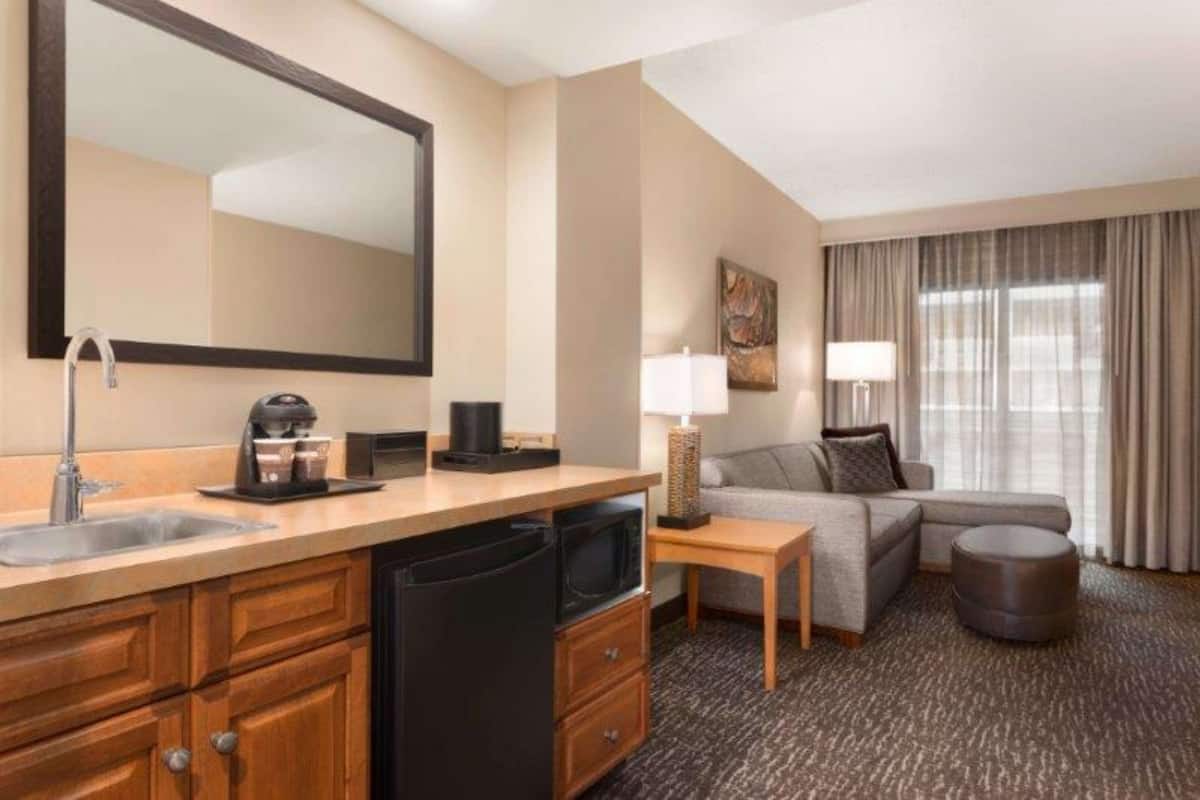 Hotelkamer met kitchenette, een bank, een lamp en een groot raam met gordijnen.
