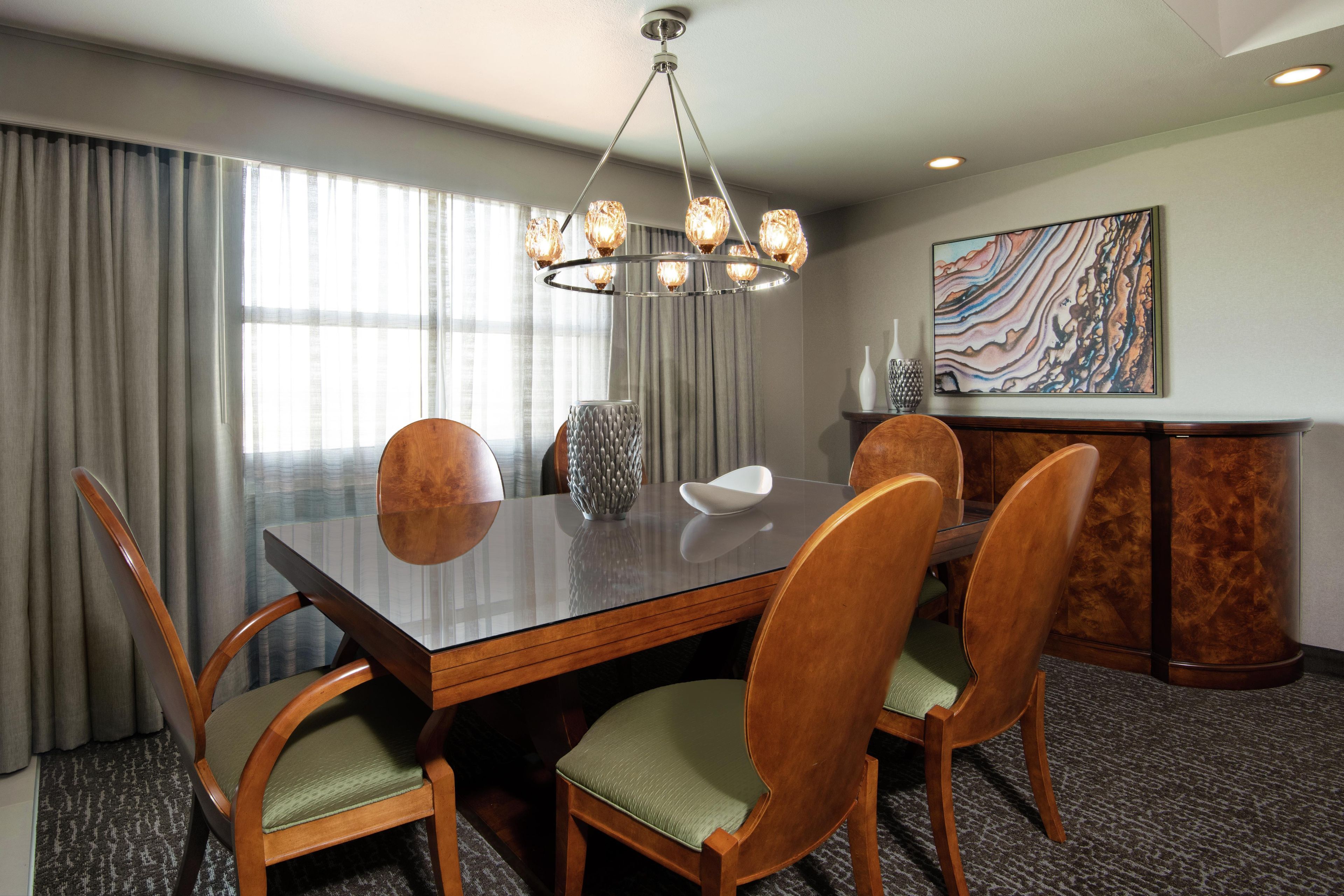 A hotel dining room with a large rectangular table, six wooden chairs with green cushions, a modern chandelier, and a decorative vase.