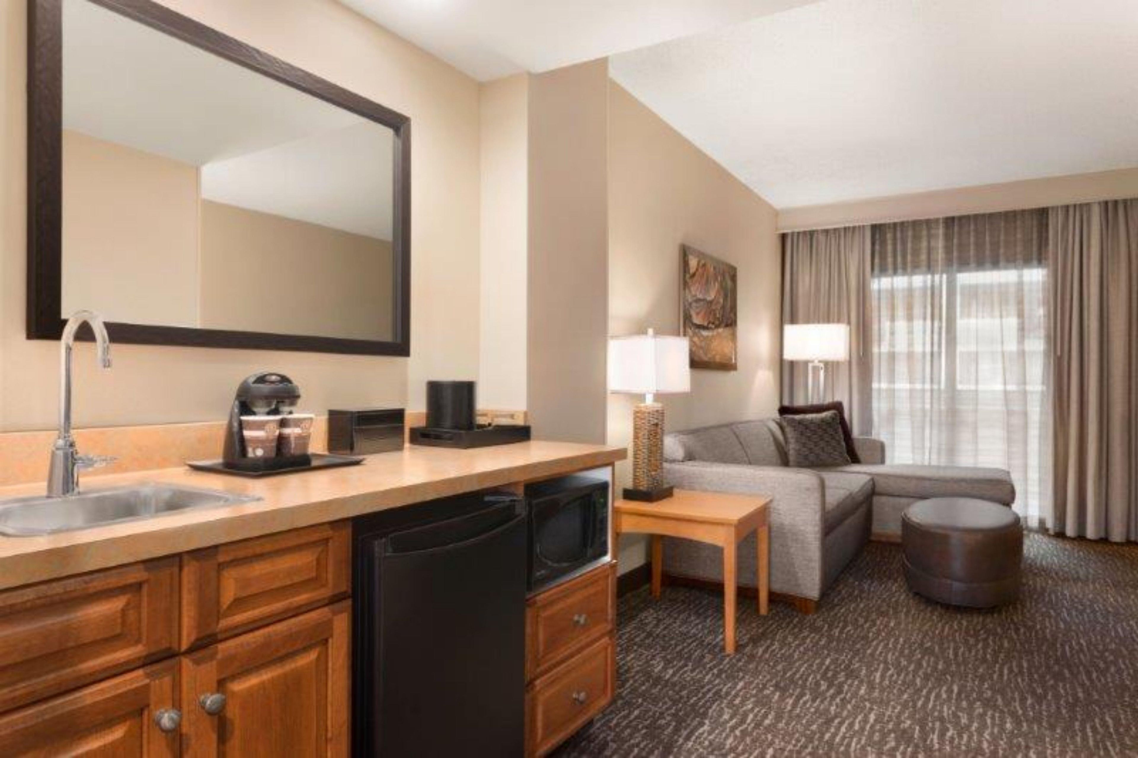 A hotel room with a kitchenette, a sofa, a lamp, and a large window with curtains.