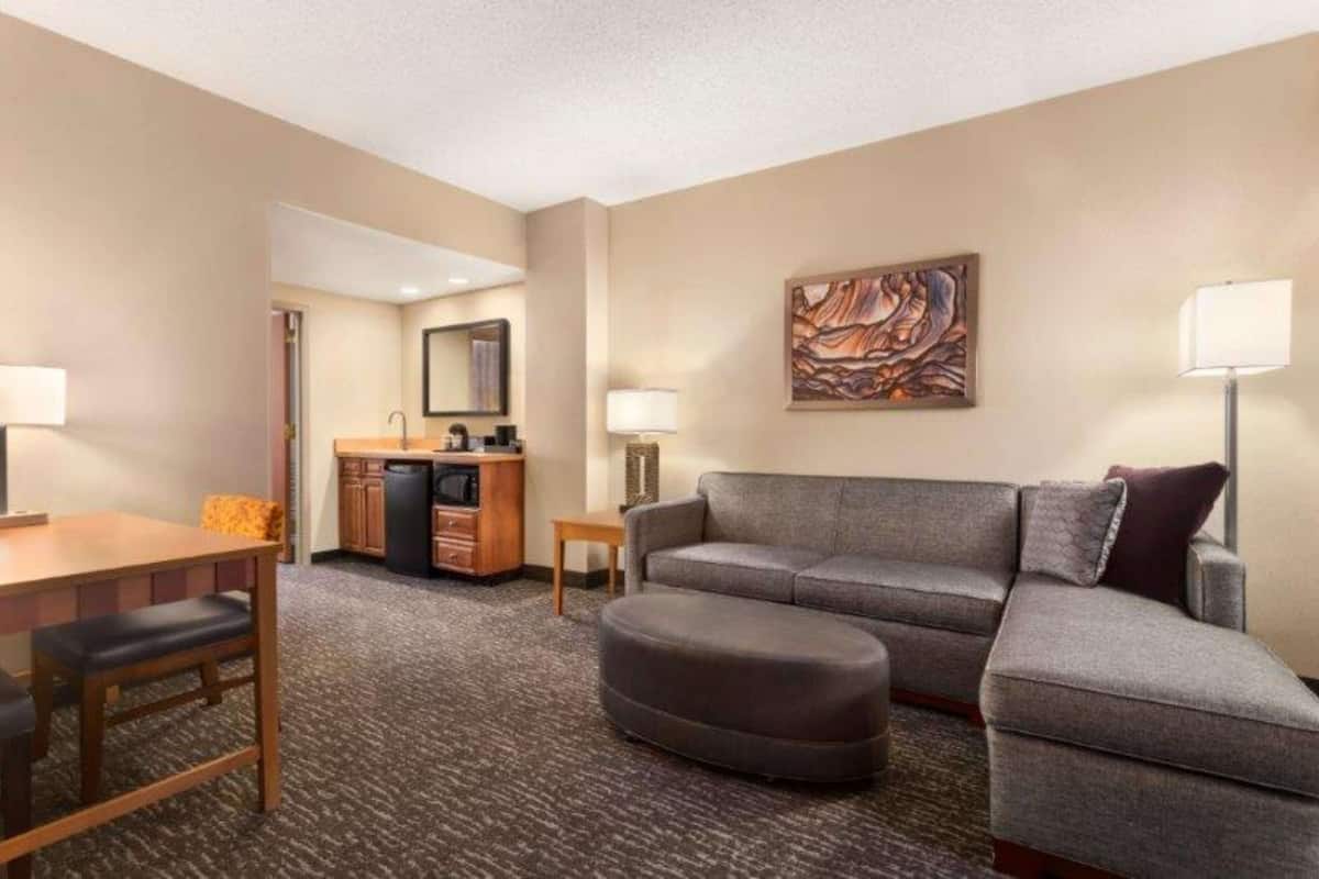 Een hotelkamer met een grijze bank, een rond poefje, een houten eettafel met stoelen, een klein bijzettafeltje en een kitchenette.