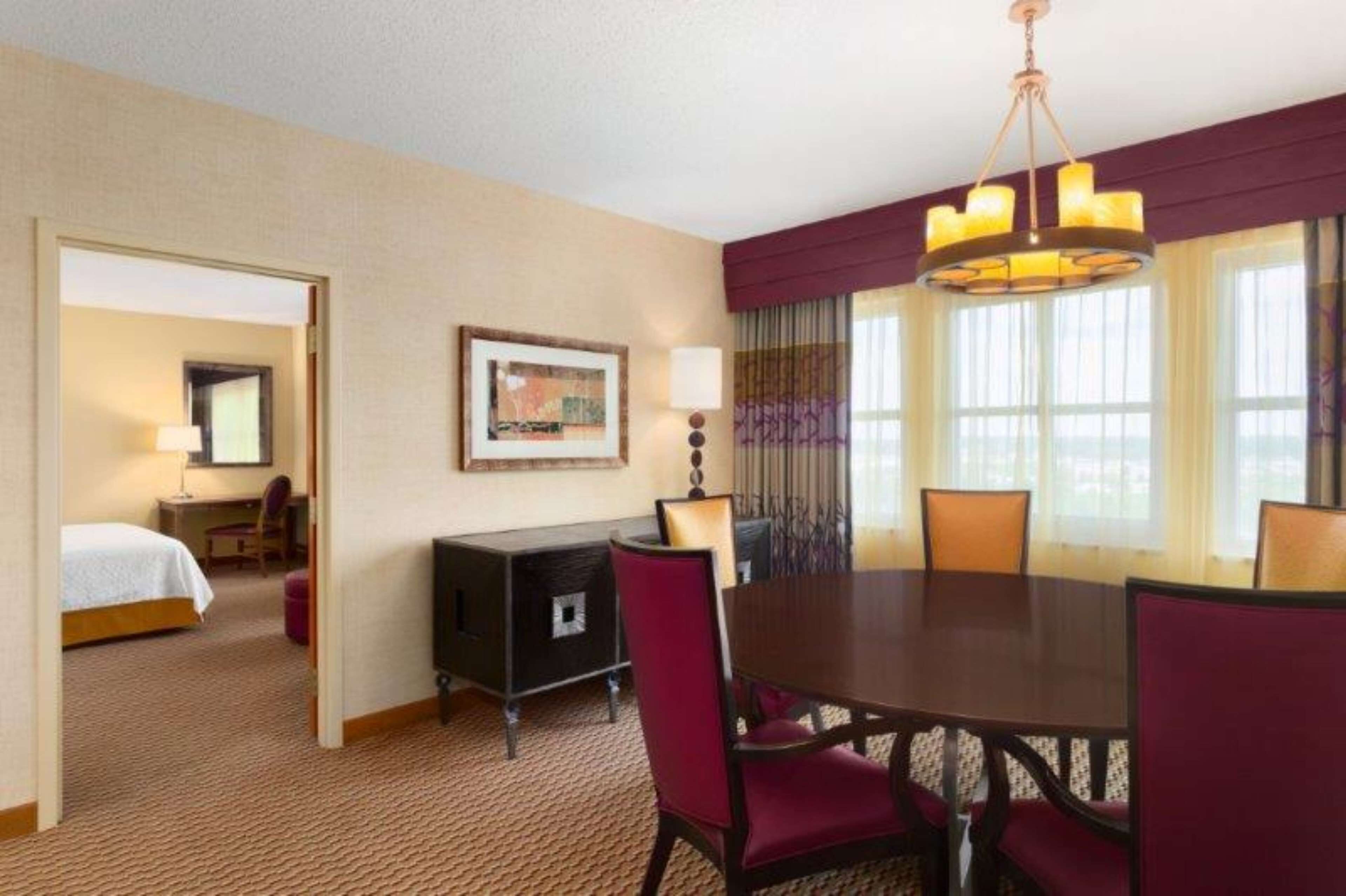 A hotel room with a bed, a desk, a dining table with chairs, a chandelier, and a large window with curtains.