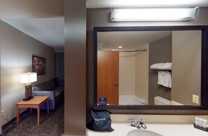 Une chambre d’hôtel avec un canapé, une table avec une lampe, un grand miroir, une salle de bain avec une baignoire et un lavabo équipé de produits de toilette.