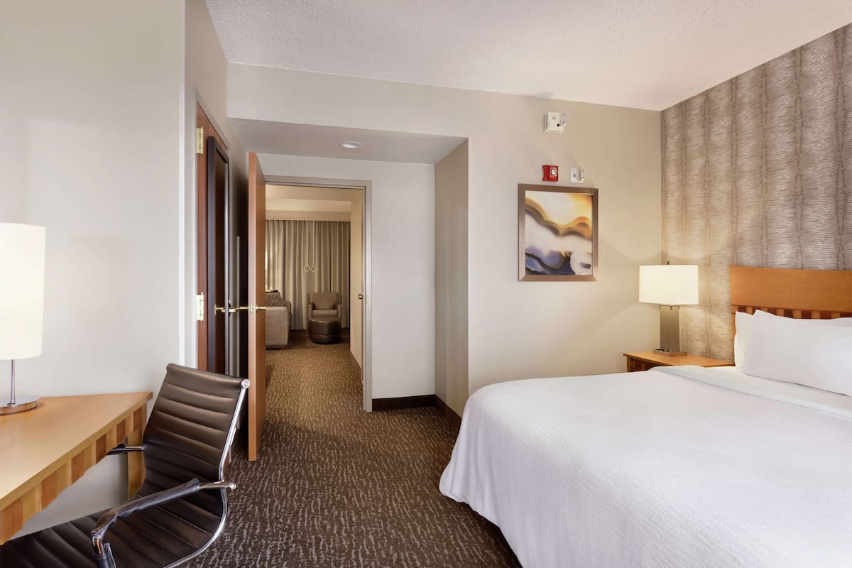 Een hotelkamer met een bed, een bureau met een stoel, een nachtkastje, een schilderij aan de muur en een deur die naar een andere kamer leidt.