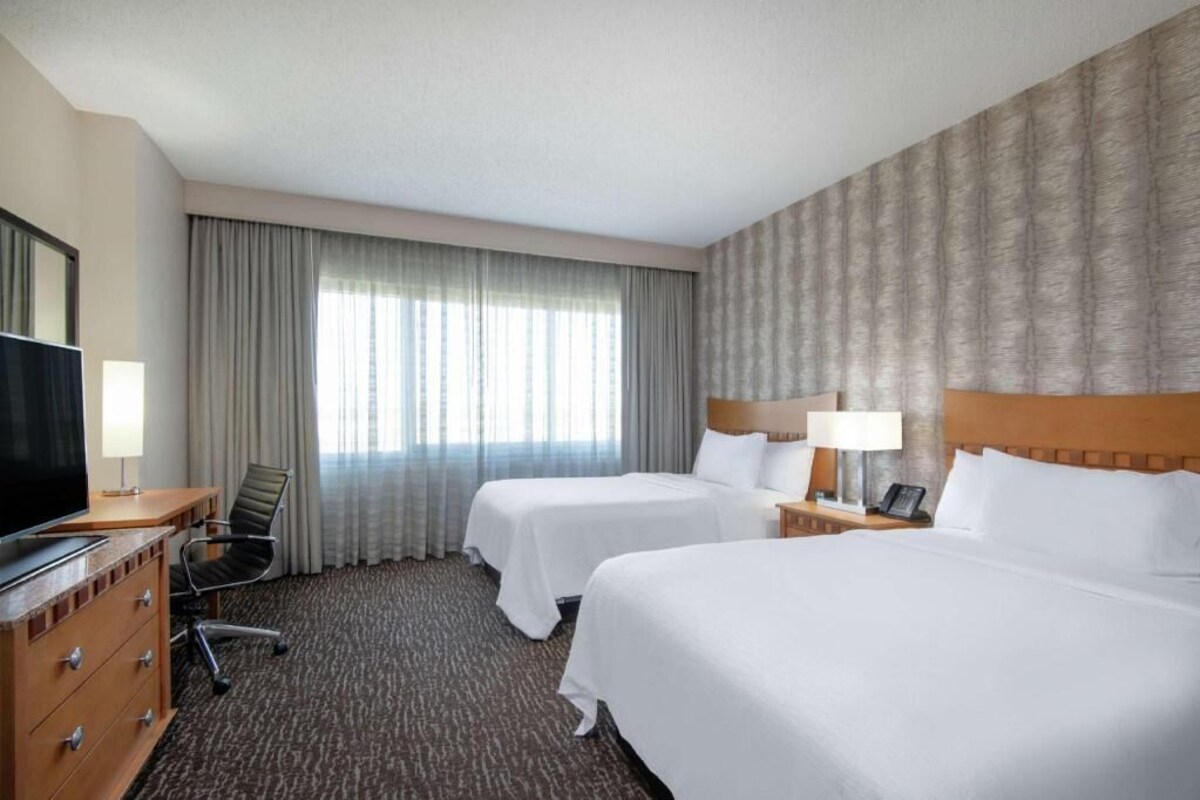A hotel room with two beds, a desk with a TV, a chair, and a large window with curtains.