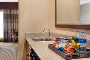 Une cuisine d’hôtel dotée d’un plan de travail en granit, d’un évier en acier inoxydable et d’un plateau contenant des collations et des bouteilles d’eau.