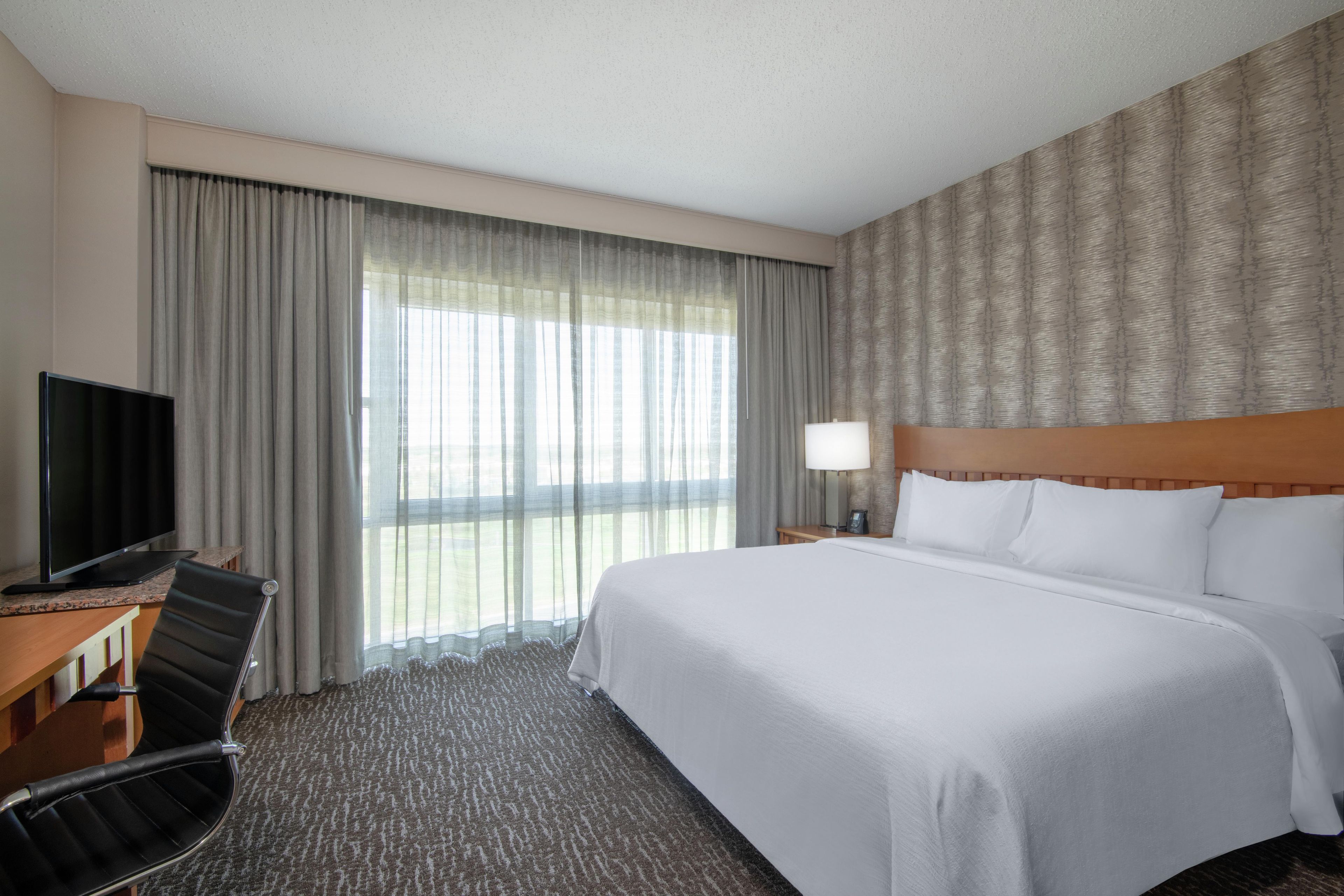 A hotel room with a bed, a desk with a television, a chair, a lamp, and a window with curtains.