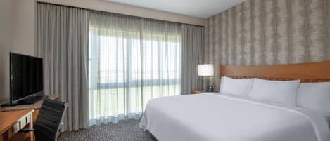 Habitación de hotel con cama, escritorio con televisor, una silla, una lámpara y ventana con cortinas.