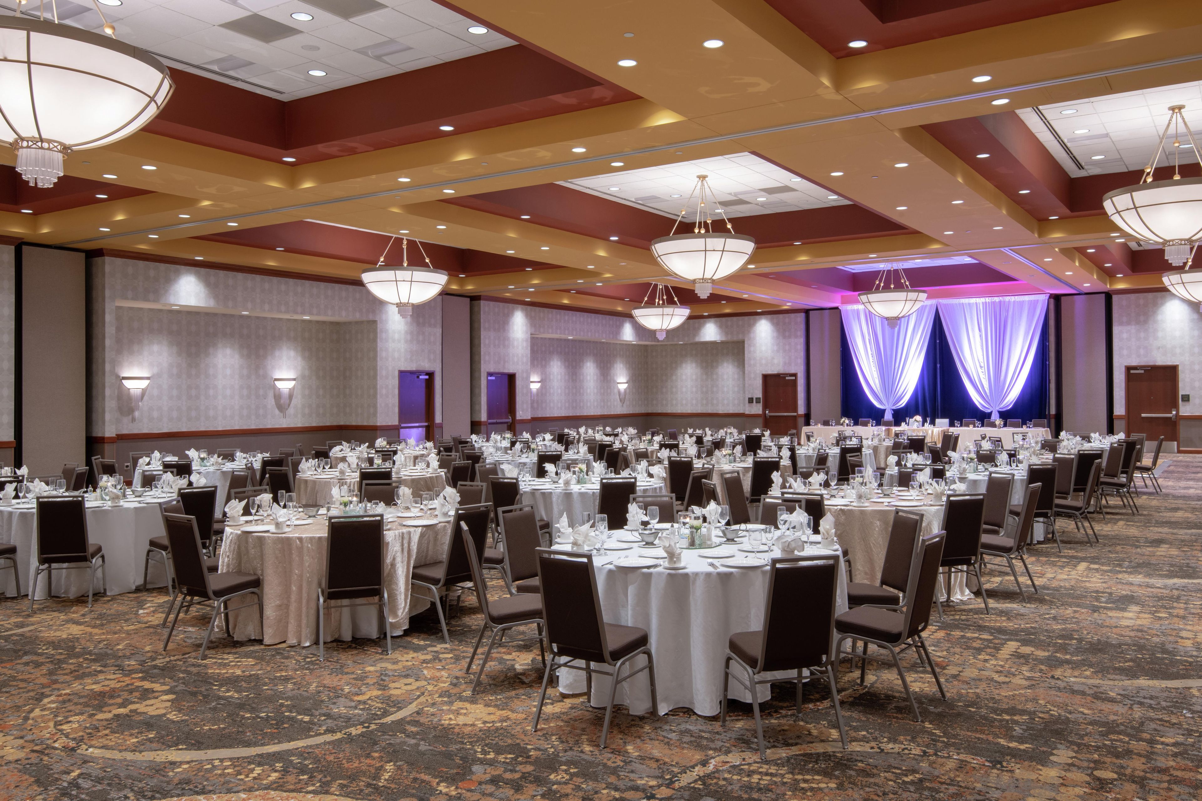 Un salón de banquetes con mesas redondas dispuestas para cenar, candelabros y un escenario con cortinas.