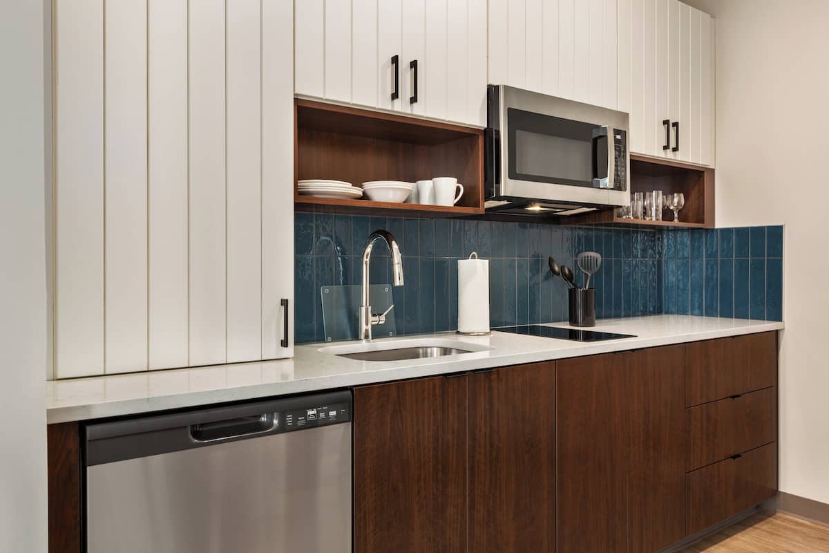 A modern kitchen with wooden cabinets, a stainless steel dishwasher, a microwave, and a blue tiled backsplash.