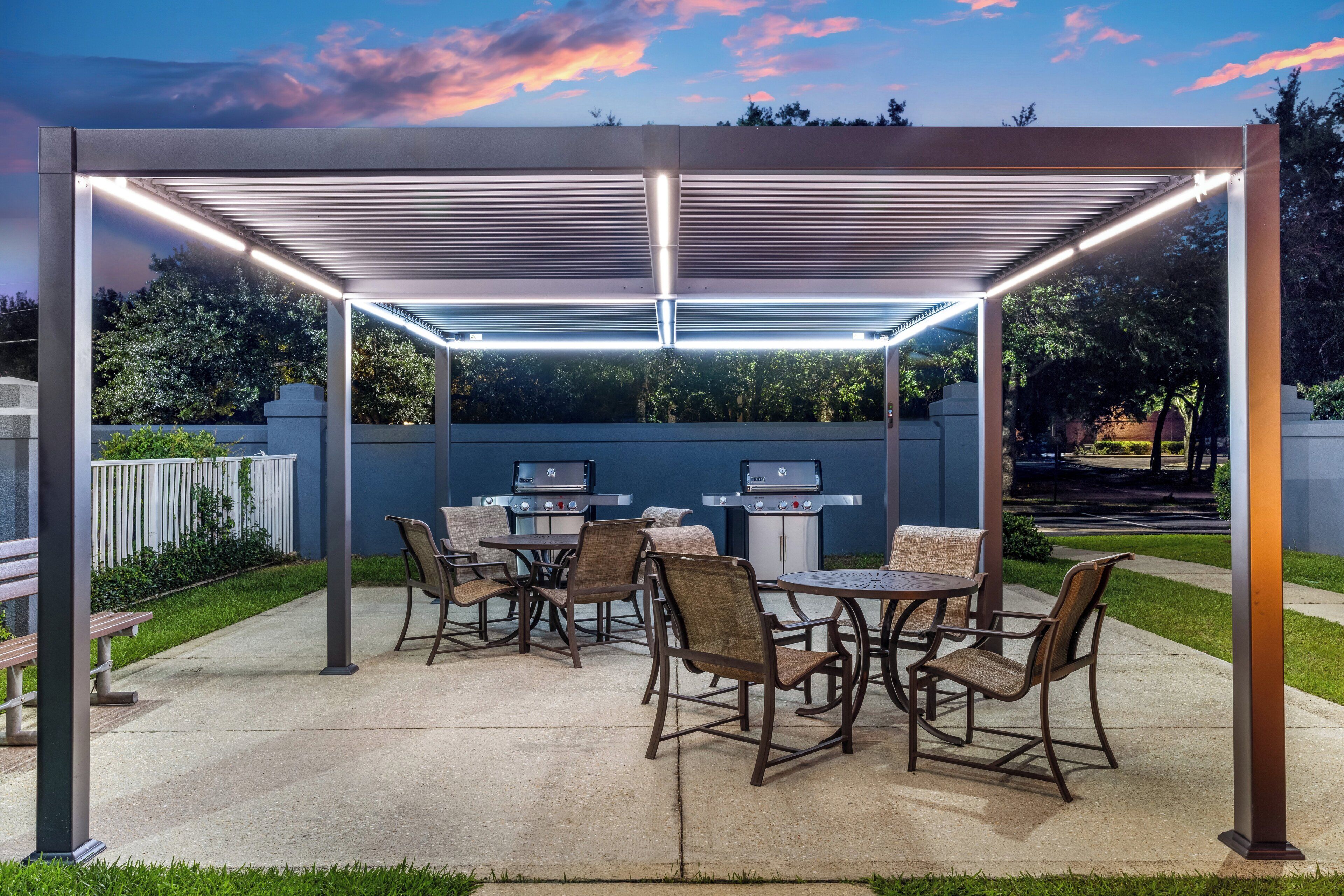 Un patio exterior con pérgola, mesas, sillas y parrillas de barbacoa.