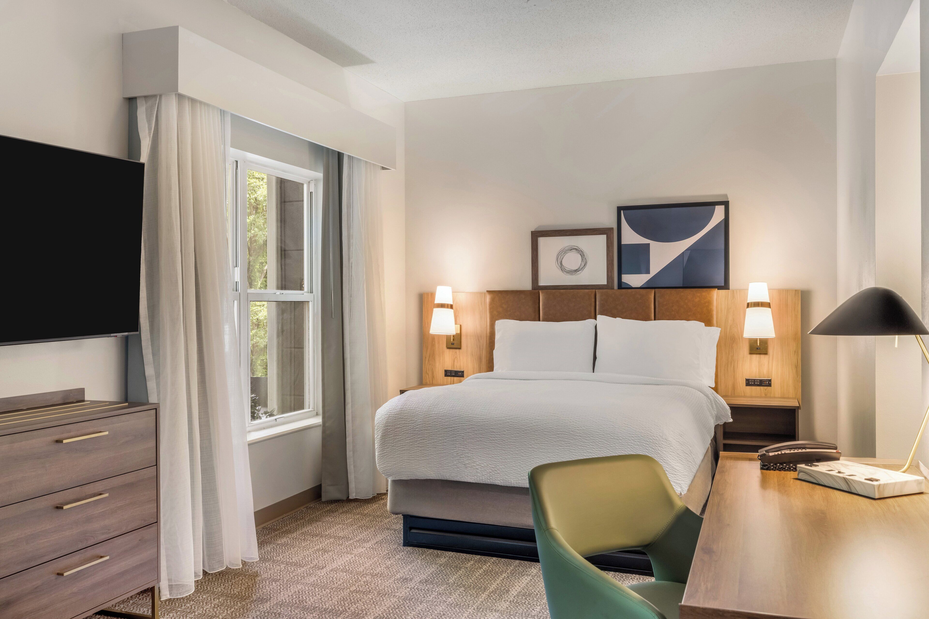A hotel room with a bed, bedside lamps, a desk, a chair, a TV, and a window with curtains.