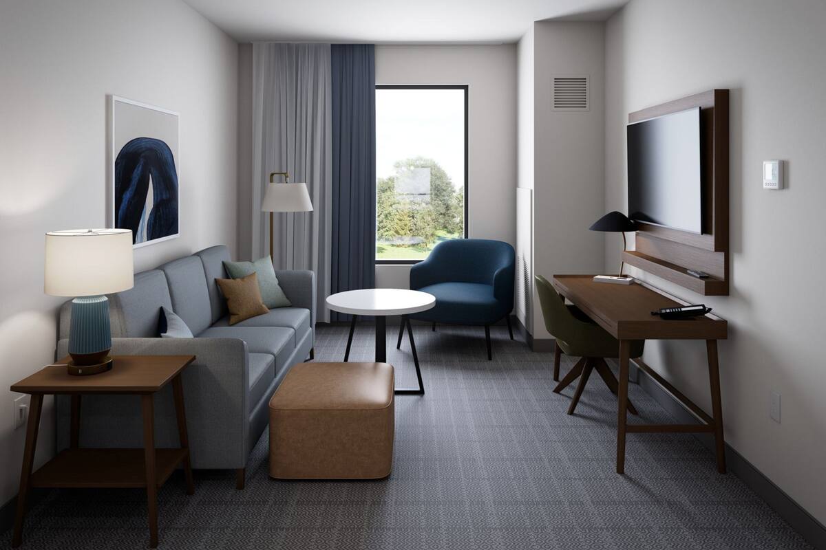 A modern hotel room with a sofa, a round table, a blue armchair, a desk, a TV, and a large window with curtains.