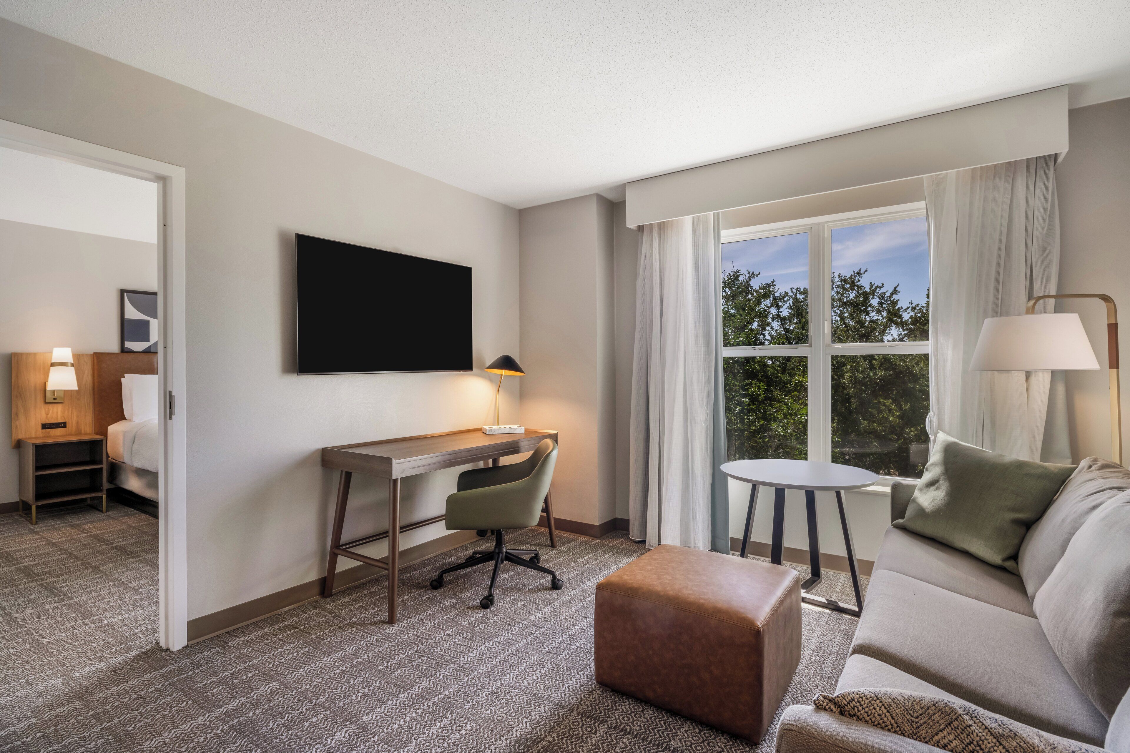 Une chambre d’hôtel équipée d’un bureau, d’une chaise, d’un canapé, d’un pouf et d’une grande fenêtre avec des rideaux.