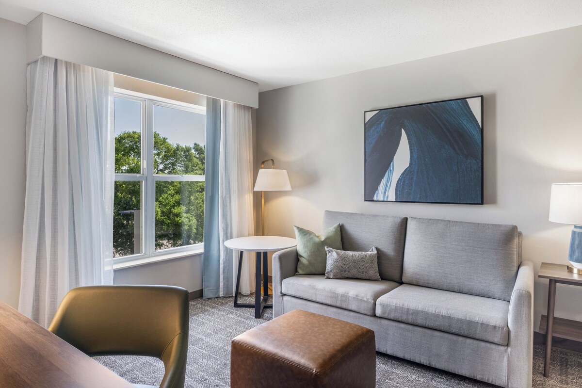 Ein modernes Hotelzimmer mit grauem Sofa, einem kleinen runden Tisch, einem braunen Hocker und einem großen Fenster mit Vorhängen.