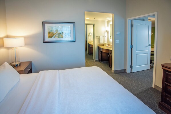 A hotel room with a large bed, a bedside table with a lamp, a framed picture on the wall, a bathroom visible through an open door, and a wooden dresser.
