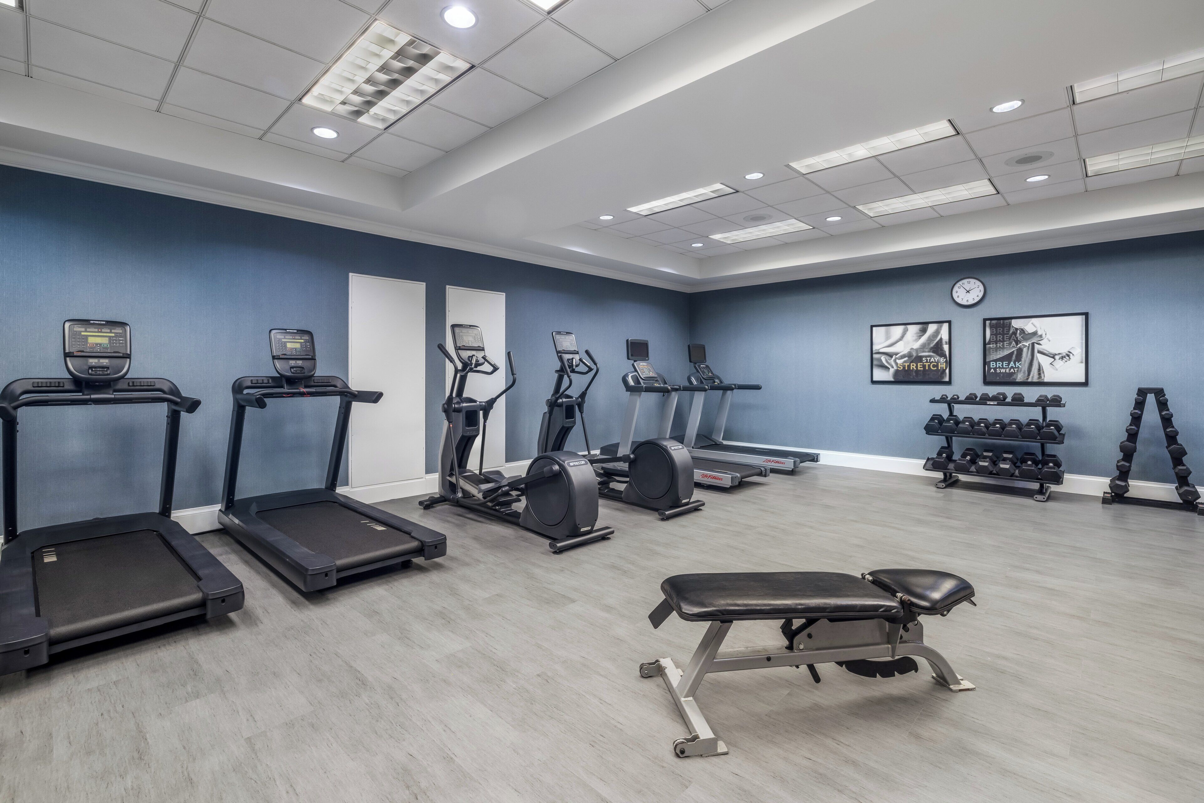 Een sportschool met loopbanden, elliptische trainers, een bank, gewichten en een klok aan de muur.