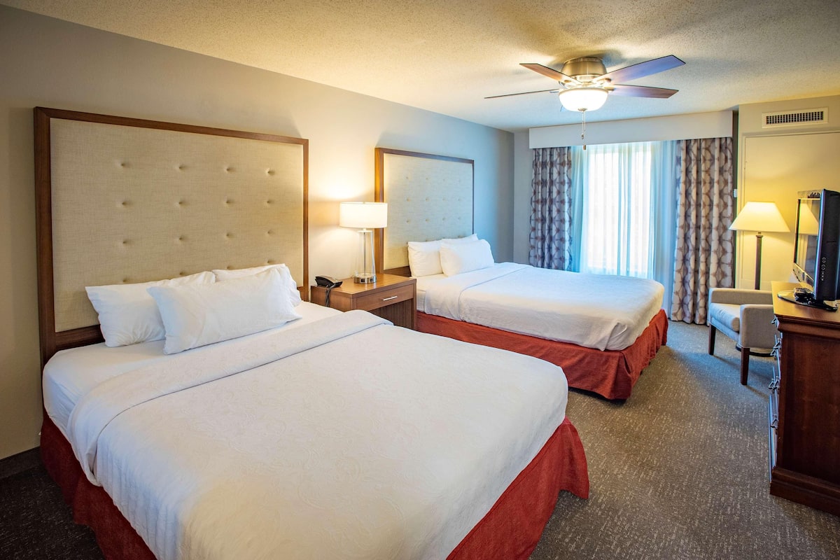 A hotel room with two beds, a ceiling fan, a desk with a lamp, and a television.