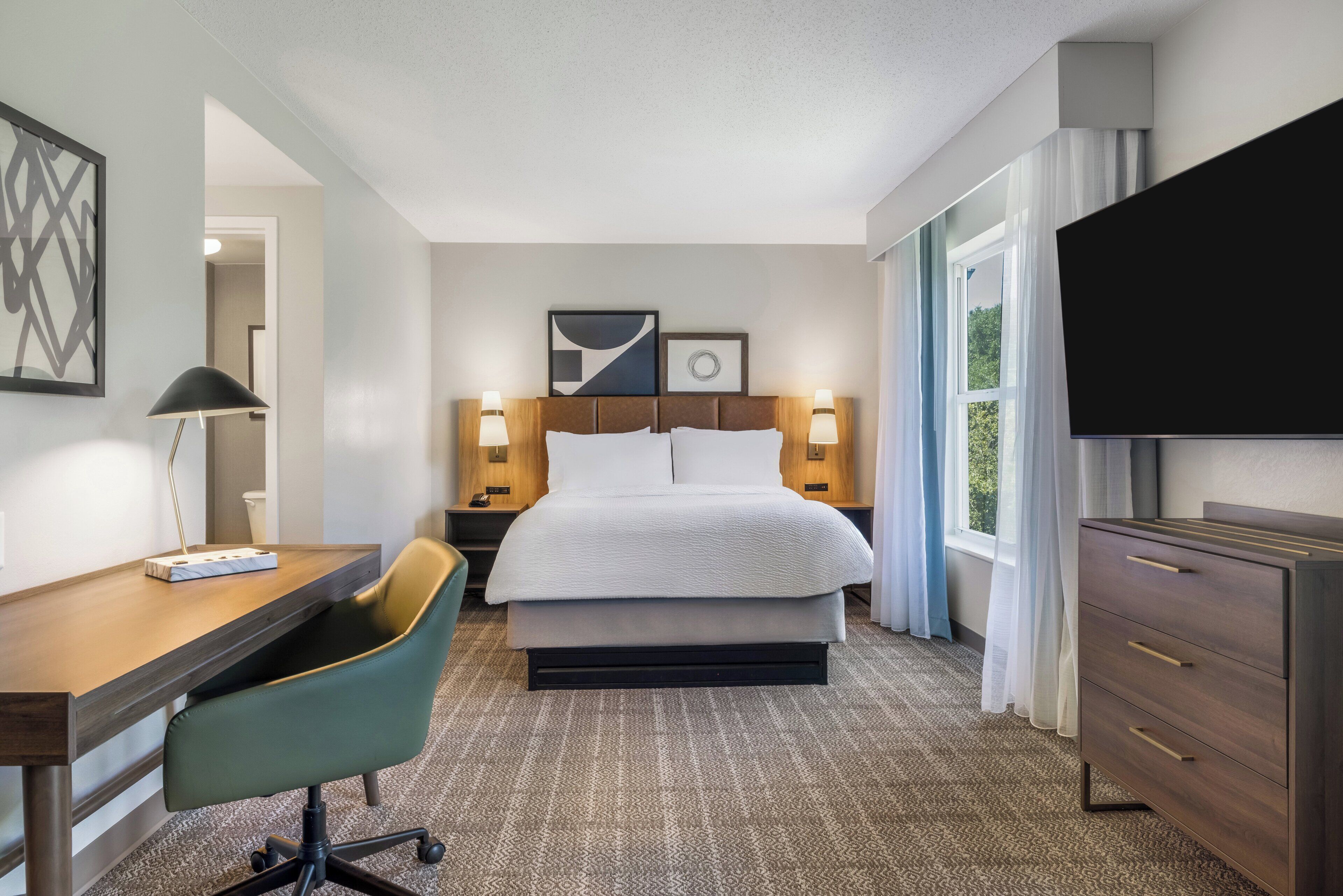 A hotel room with a bed, desk, chair, TV, and artwork on the wall.