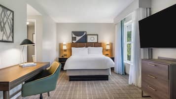 A hotel room with a bed, desk, chair, TV, and artwork on the wall.