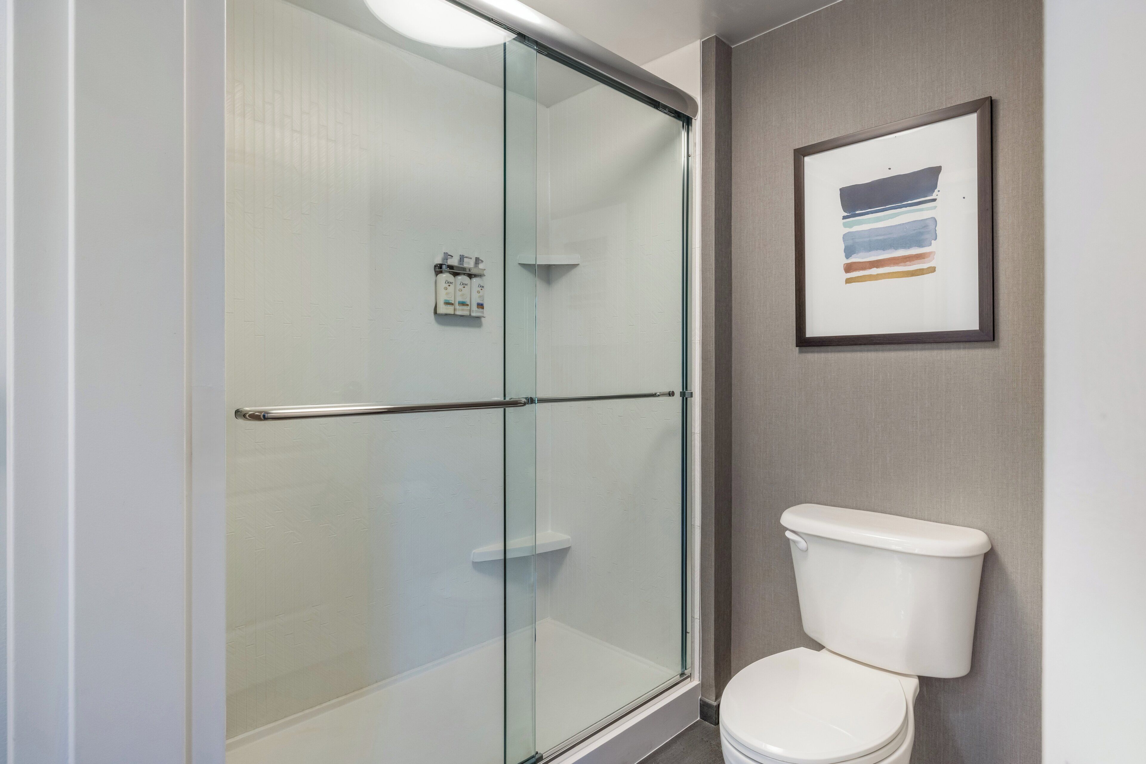 A bathroom with a glass shower enclosure, a white toilet, and a framed abstract painting on the wall.