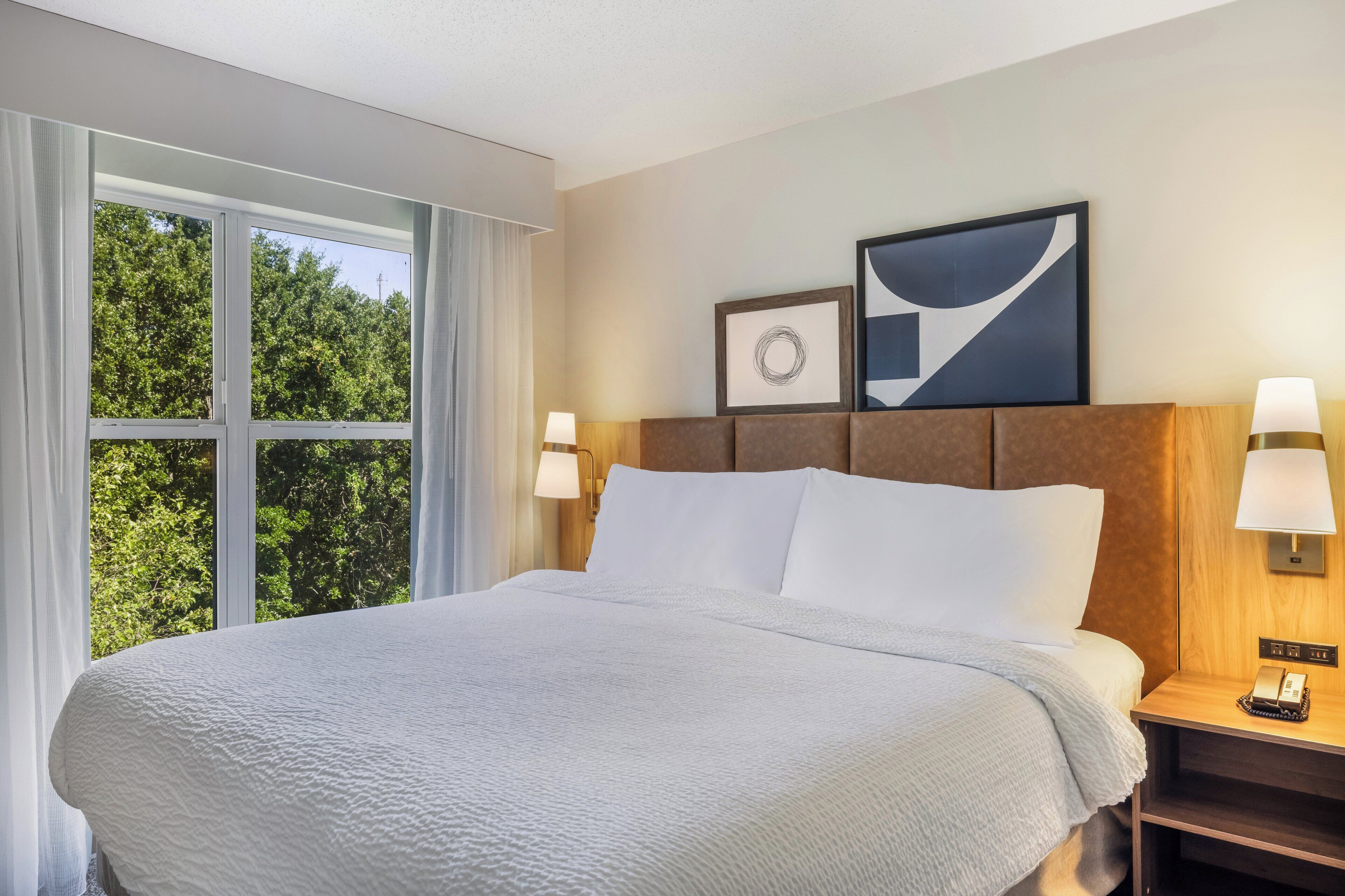 Habitación de hotel con una cama grande, dos lámparas de noche, una obra de arte abstracta enmarcada y vistas a árboles a través de una ventana.