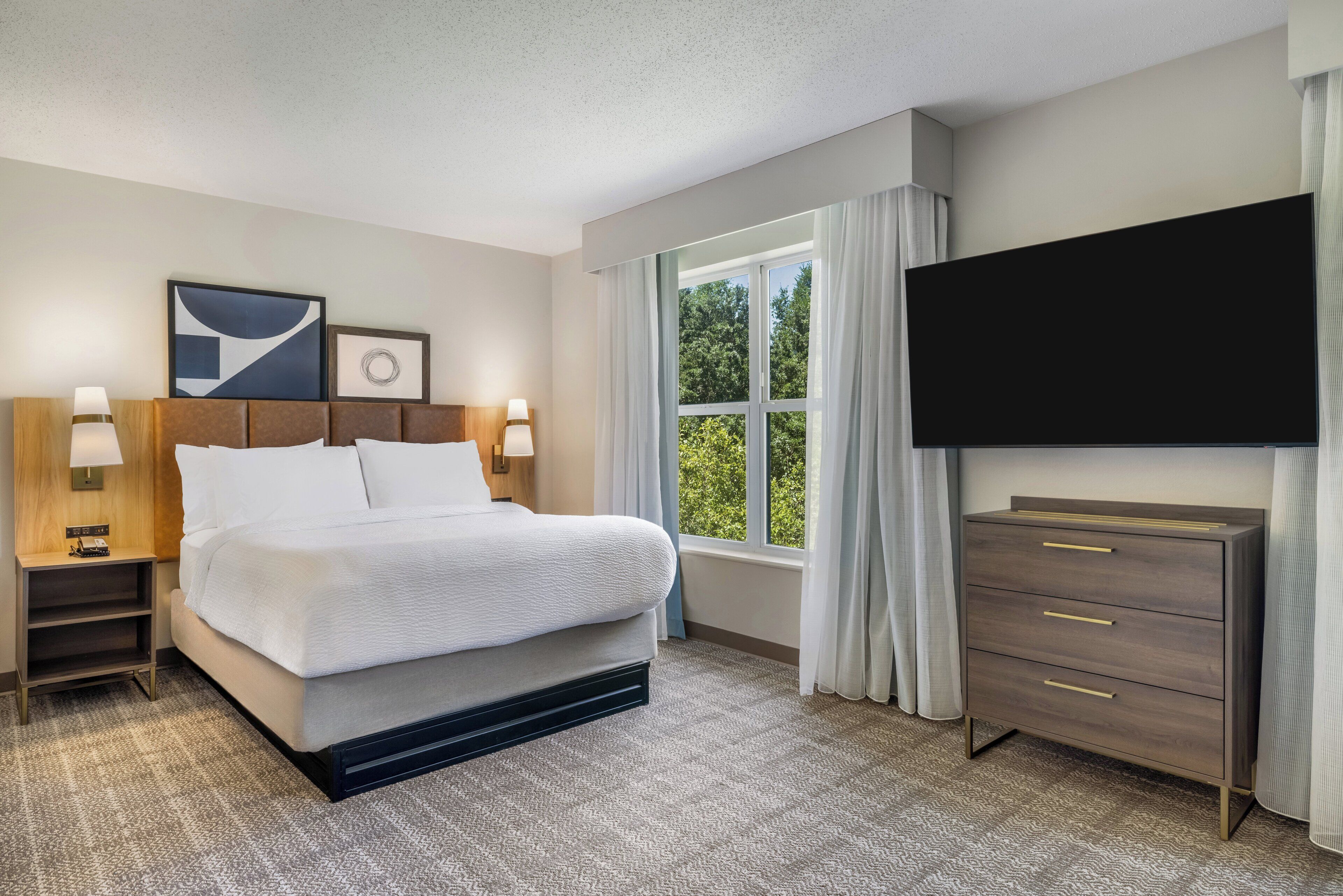 Habitación de hotel con cama, mesitas de noche, televisor, cómoda y ventana con cortinas.