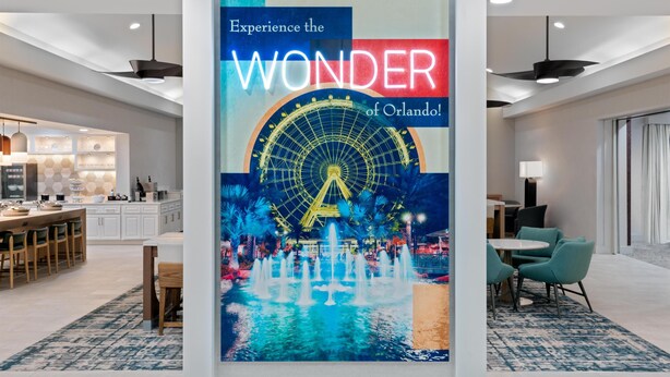 A modern hotel interior with a central column featuring a large poster of a Ferris wheel and water fountains, a dining area with a kitchen in the background, and a seating area with a view of the ocean.
