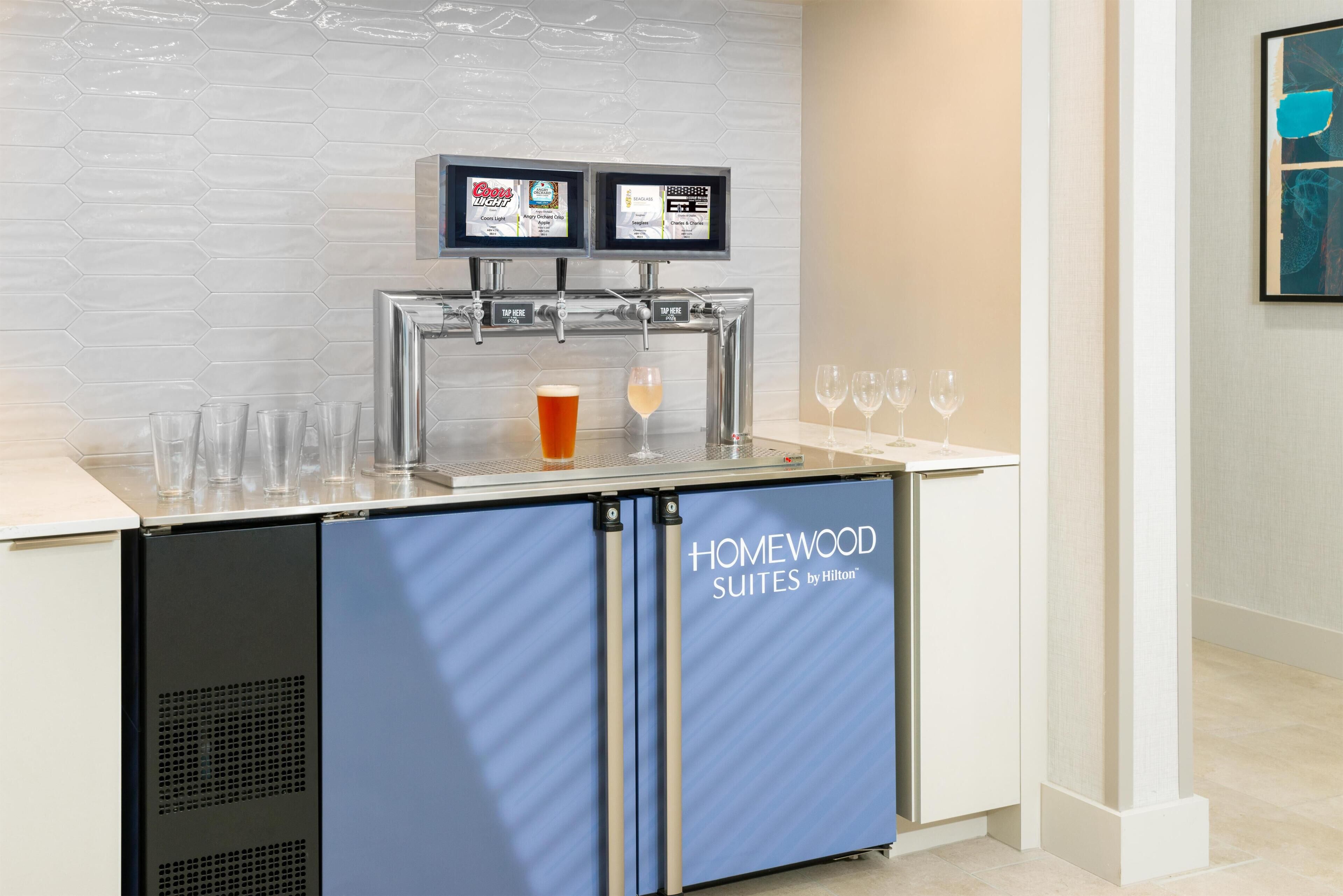 A minibar with a beverage dispenser, glasses, and a sign indicating 'HOMEWOOD SUITES by Hilton'.