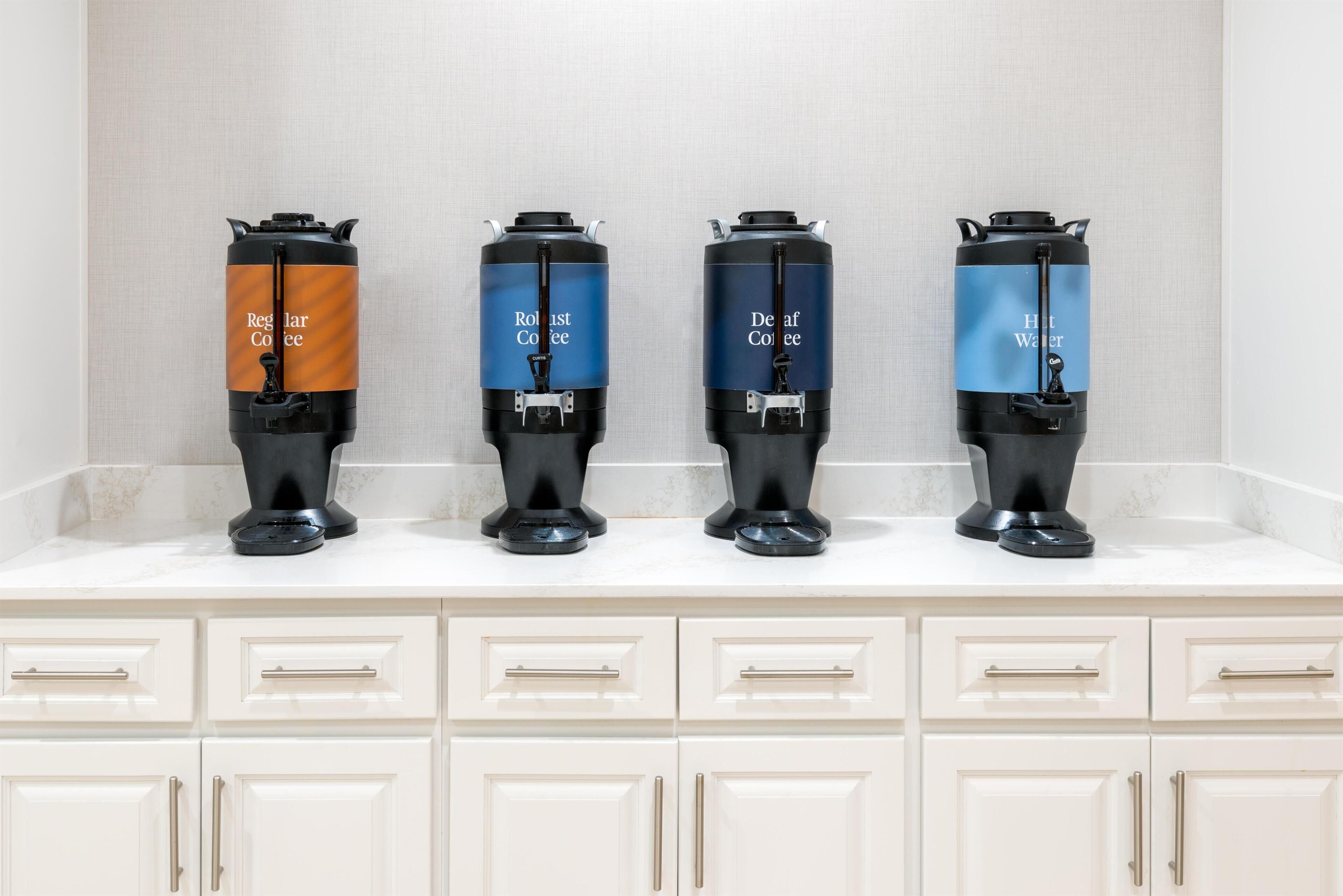 Four coffee dispensers on a countertop with a tiled backsplash and white cabinets below.