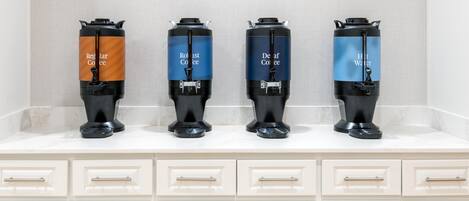 Four coffee dispensers on a countertop with a tiled backsplash and white cabinets below.