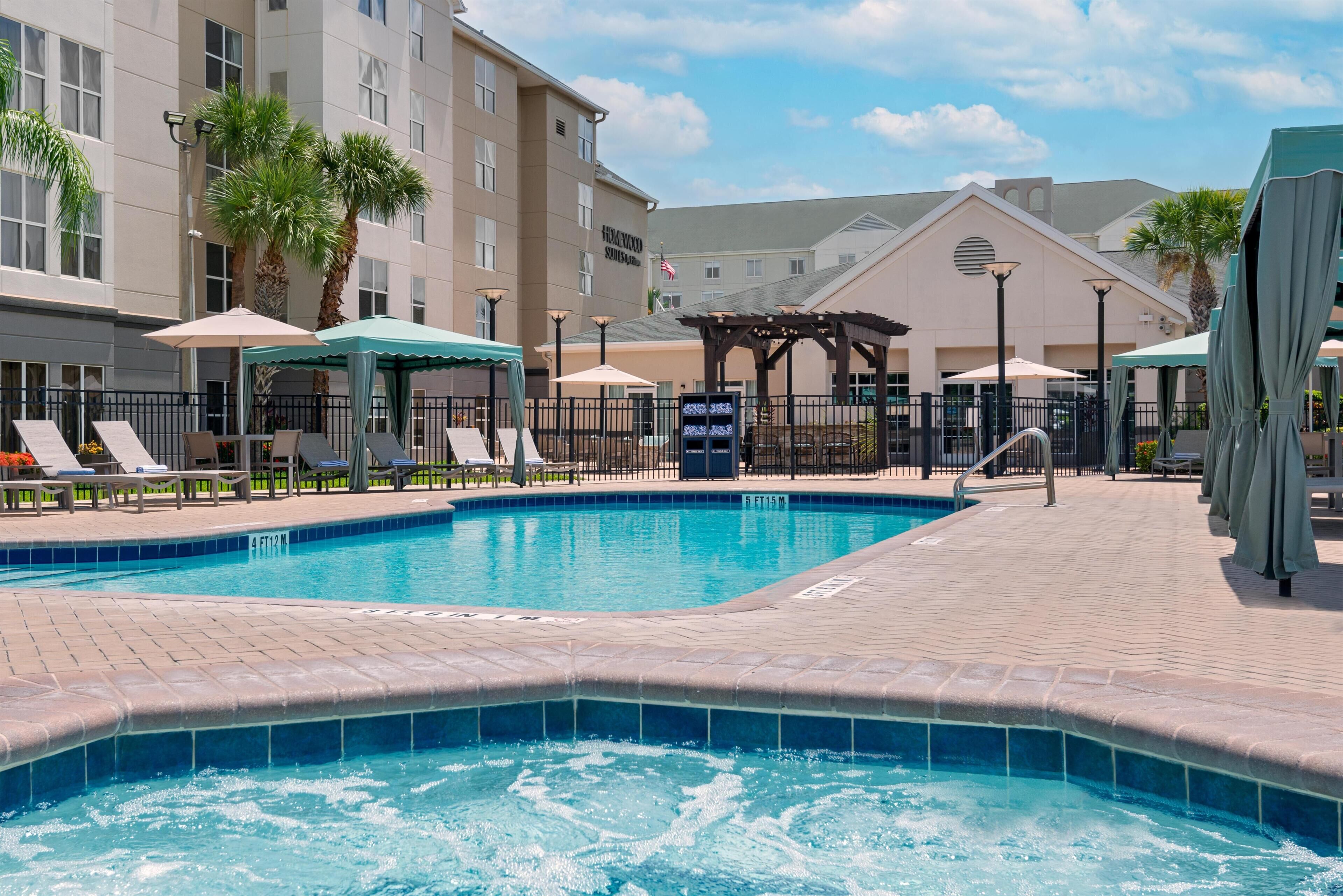 A swimming pool with lounge chairs and umbrellas, surrounded by a brick walkway and a building with a sign.