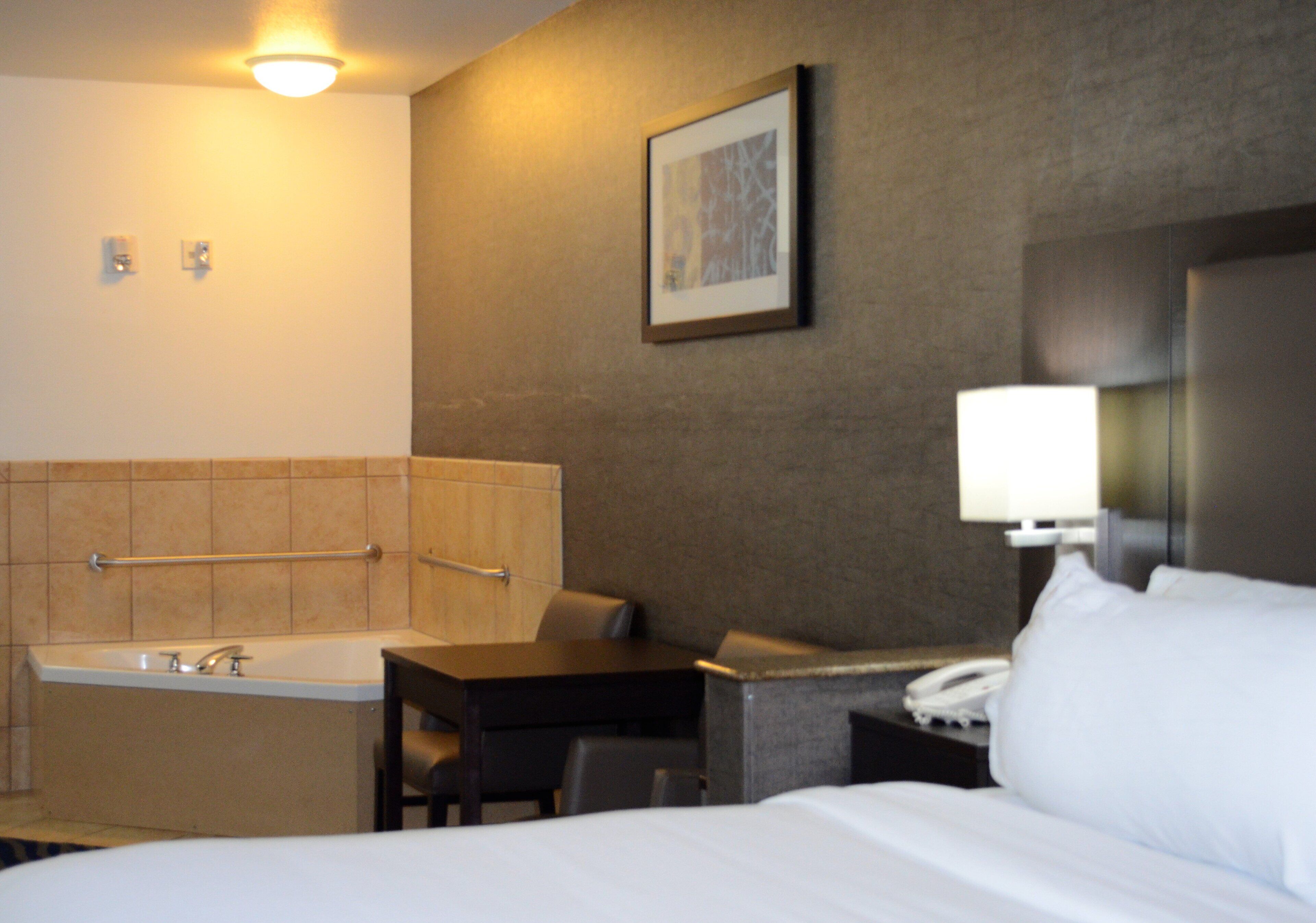 A hotel room with a bed, a desk, a chair, a telephone, a lamp, a painting, a bathtub, and a wall-mounted light fixture.
