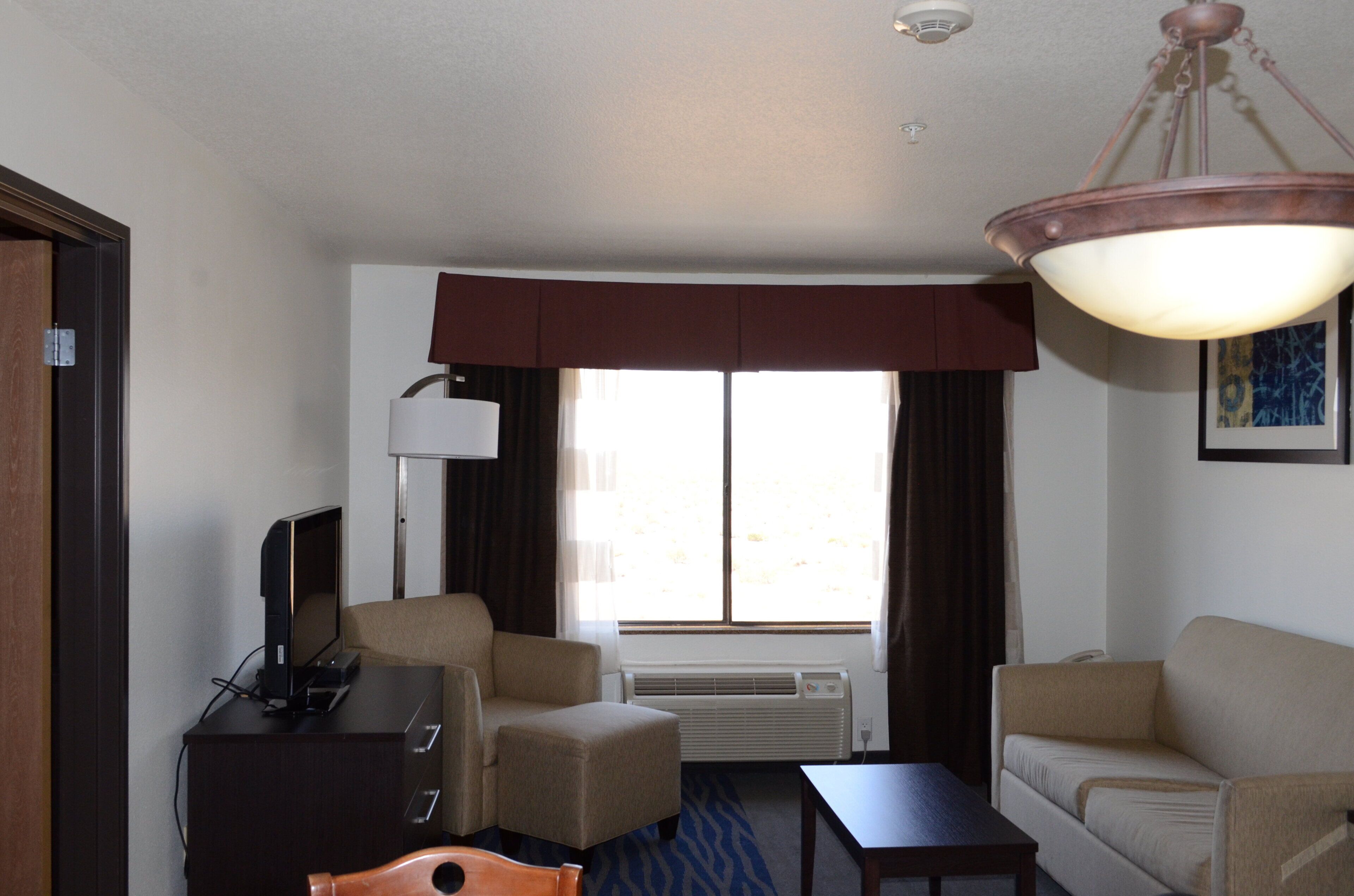 Une chambre d’hôtel équipée d’un bureau, de deux chaises, d’une télévision, d’une lampe, d’un climatiseur et d’une fenêtre avec des rideaux.