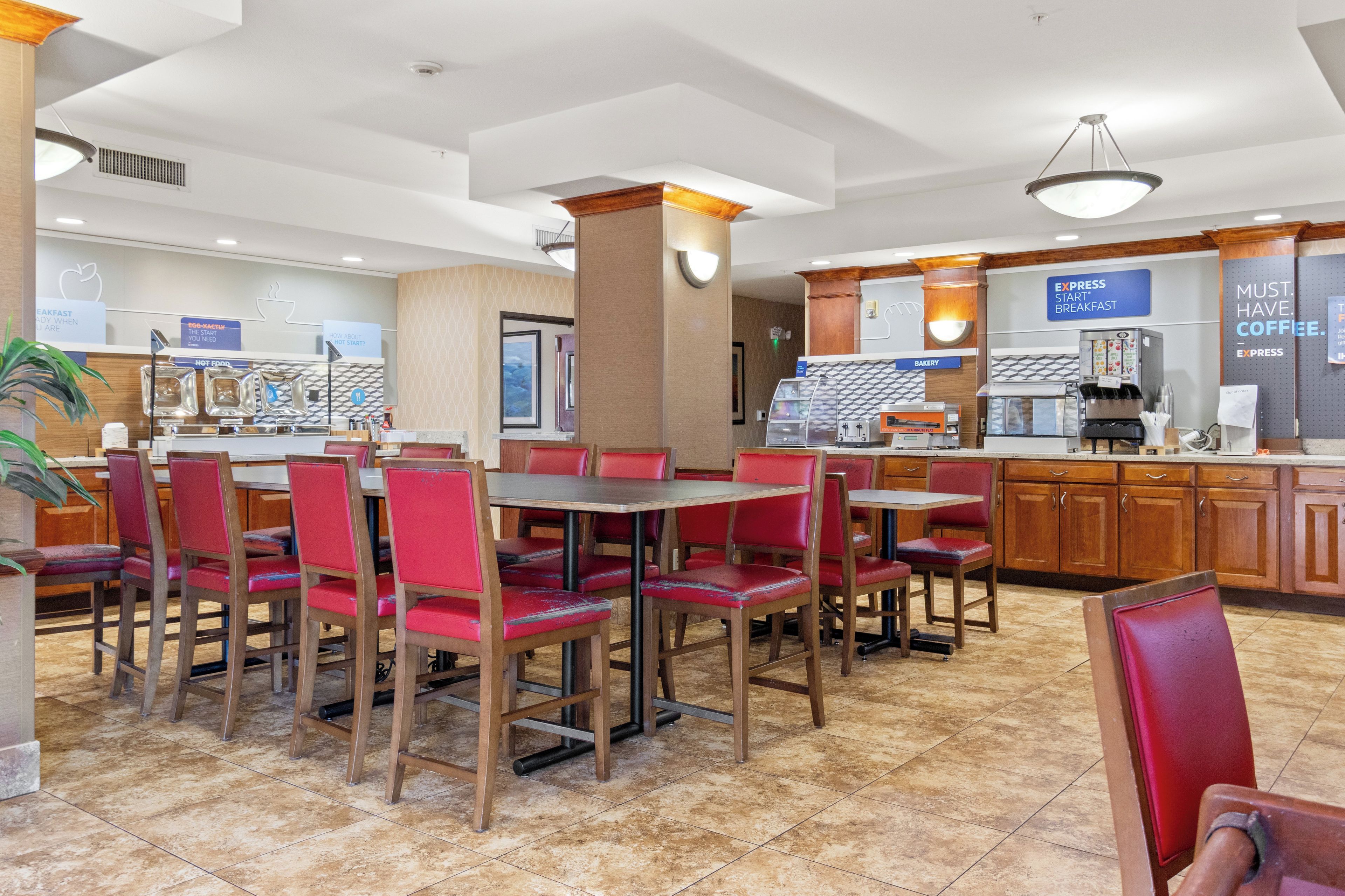 A hotel breakfast area with red upholstered chairs and tables, a coffee station, and a bakery section.