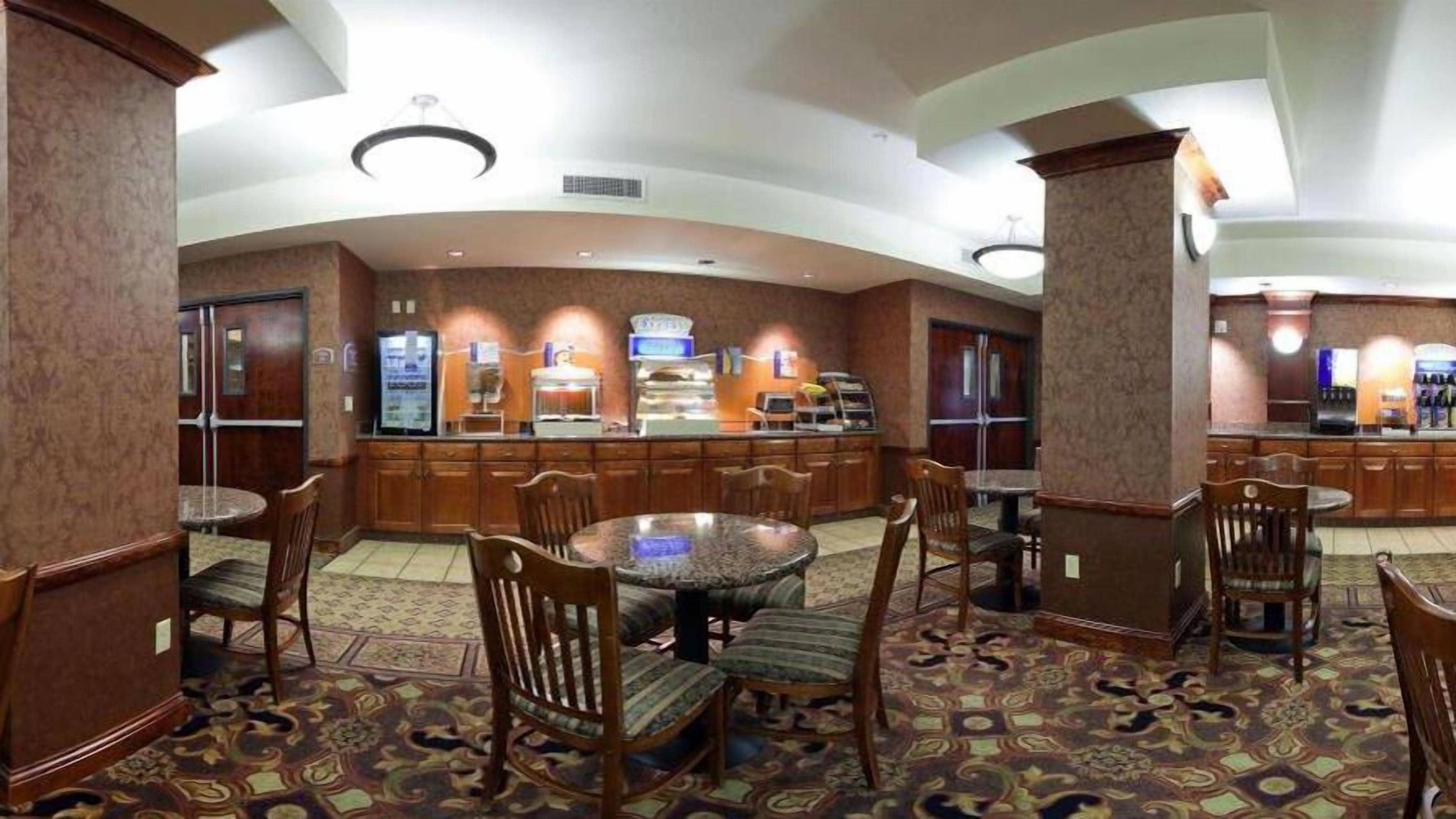 Recepció d'hotel amb una taula rodona central envoltada de cadires, zona de buffet amb barra i exposició de menjar, i zona de bar amb seients.
