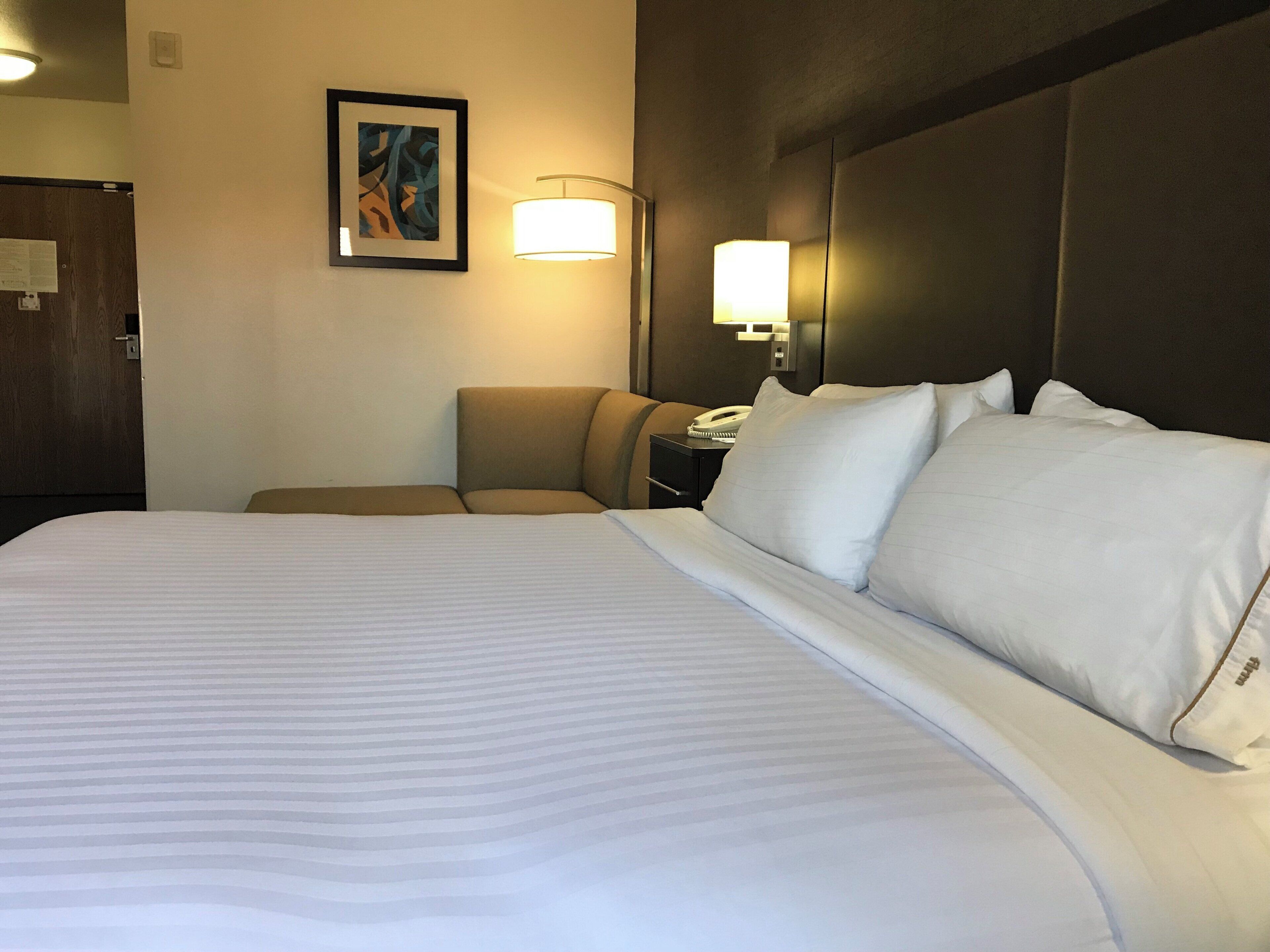 Une chambre d’hôtel avec un grand lit, une table de chevet avec une lampe, une chaise et un tableau encadré au mur.
