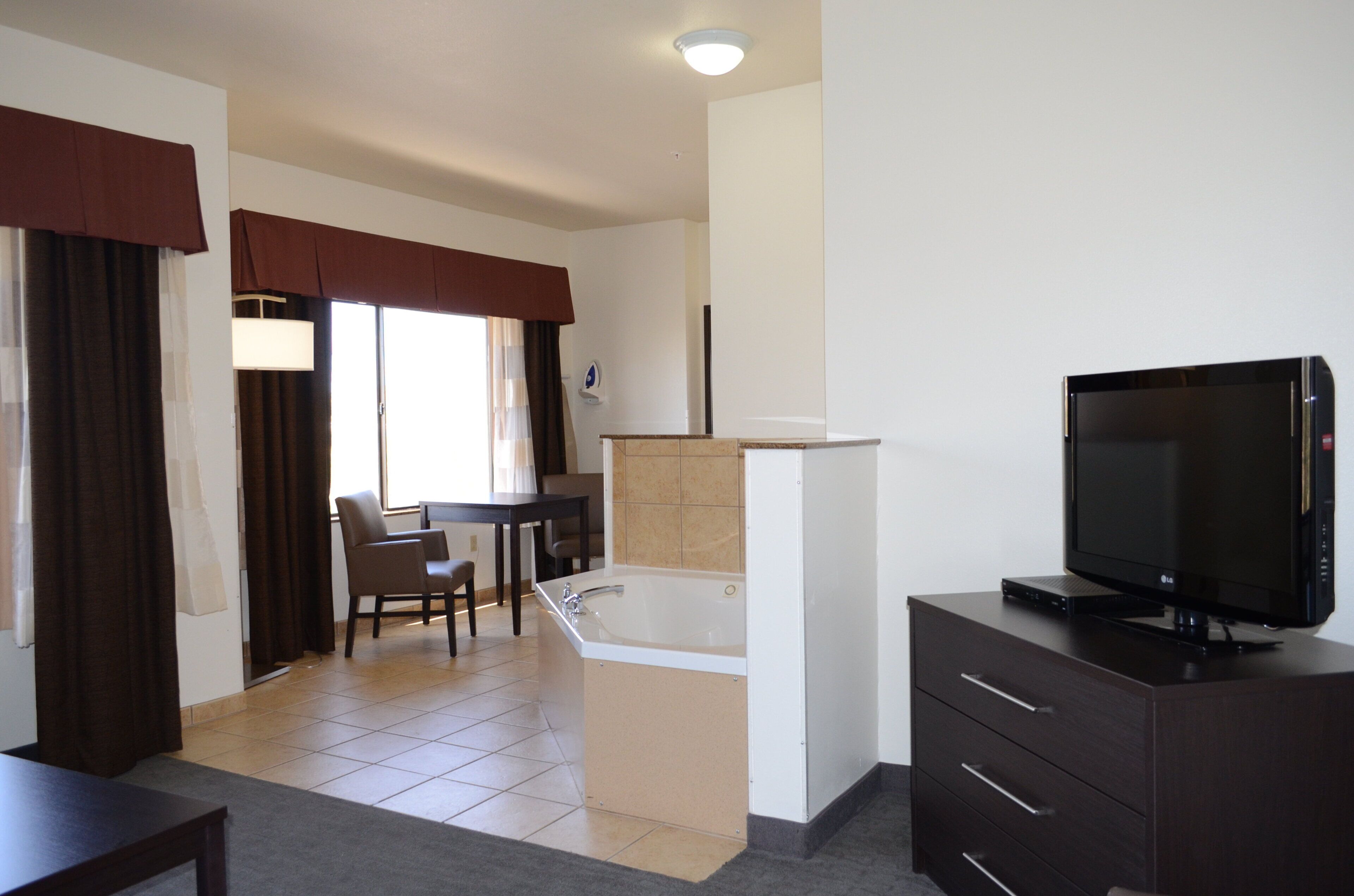 A hotel room with a television, a small table, a chair, a bathtub, and a window with curtains.