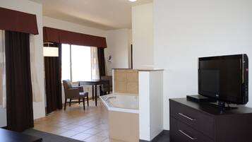 A hotel room with a television, a small table, a chair, a bathtub, and a window with curtains.