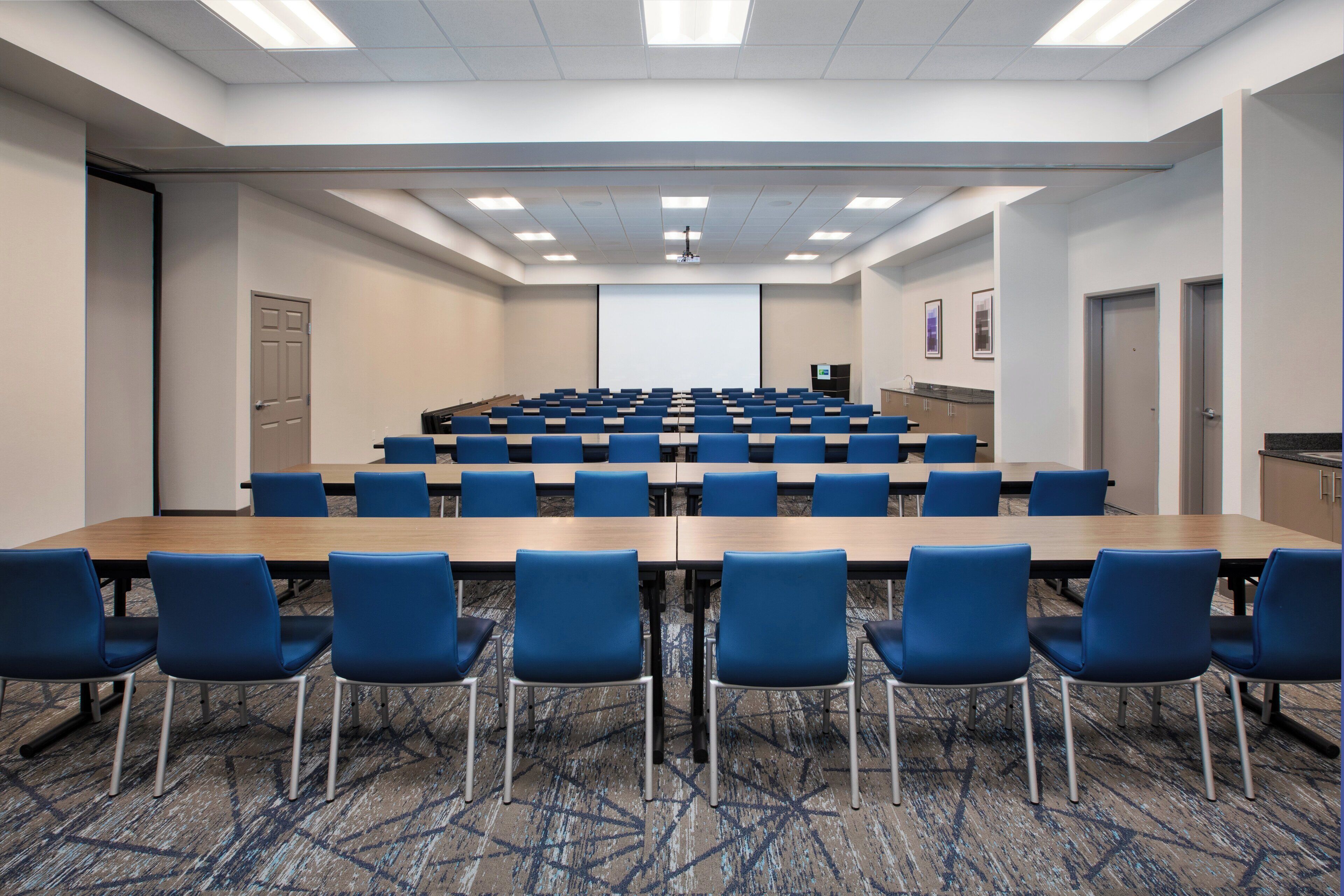 Una sala conferenze con file di sedie blu, un tavolo grande e uno schermo per proiezioni.