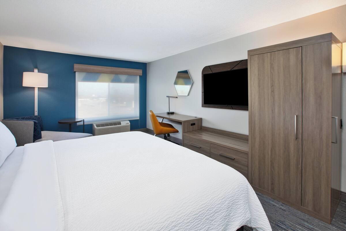 A hotel room with a large bed, a desk with a chair, a wardrobe, a TV, and a window with blinds.