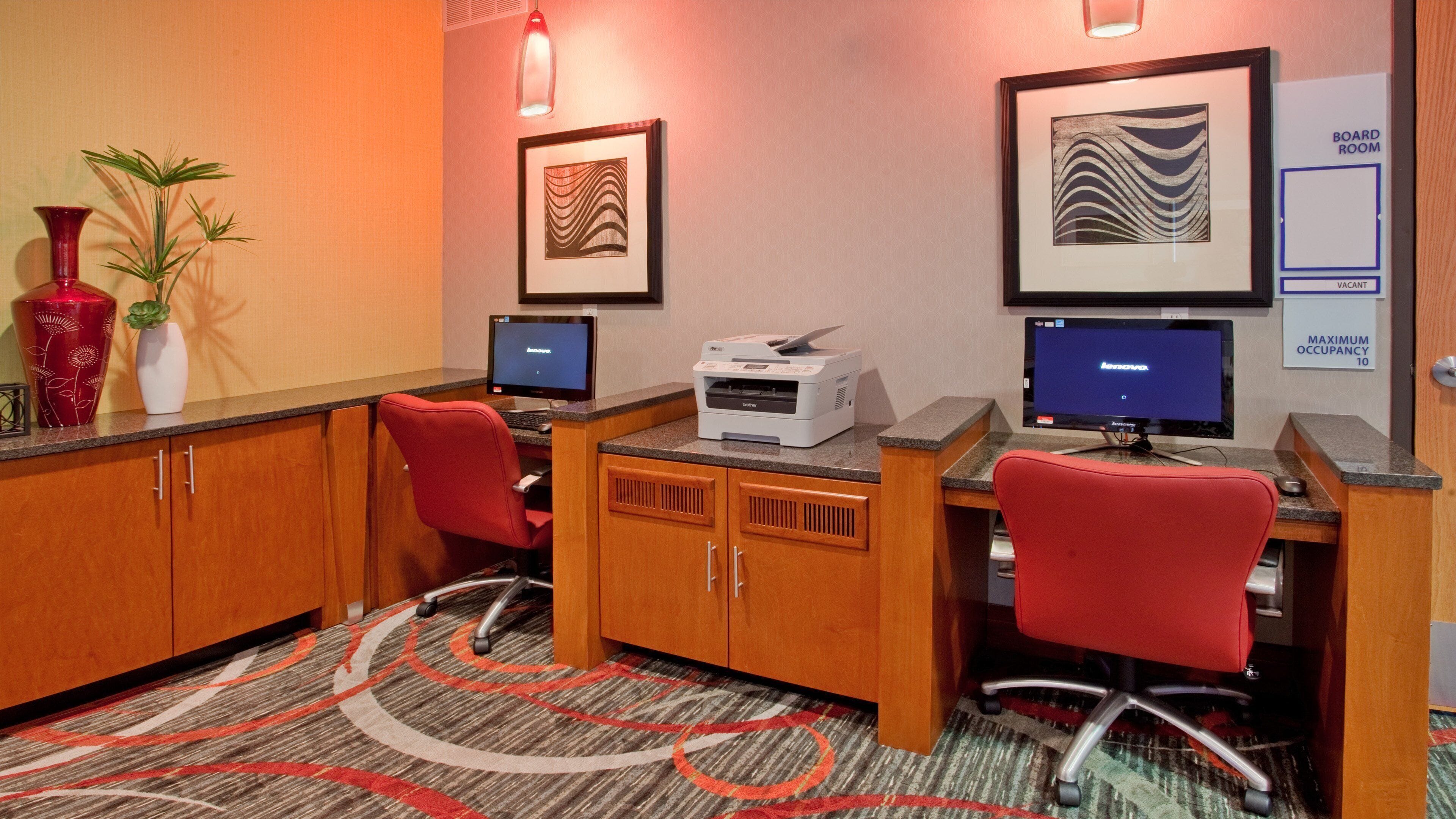 A hotel room with two desks, each with a computer and a printer, and a large framed artwork on the wall.