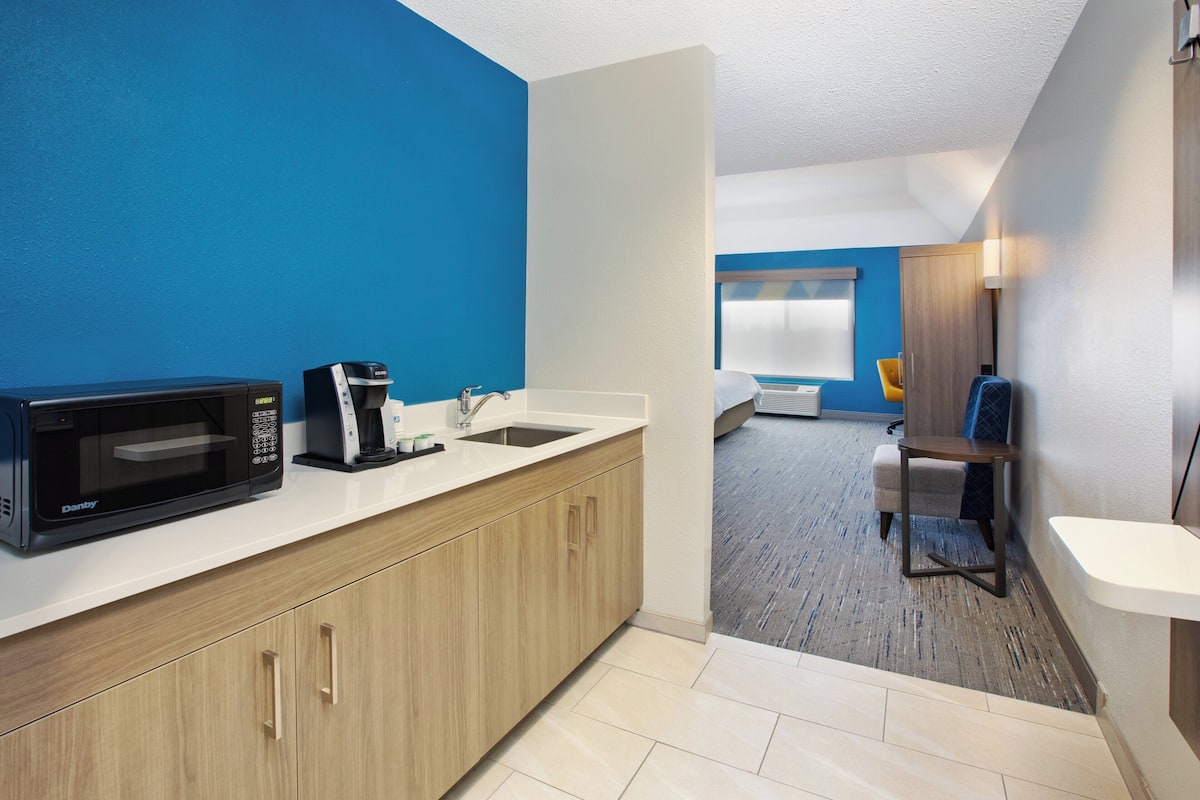A compact hotel room with a kitchenette, a bed, a small table, and a chair.