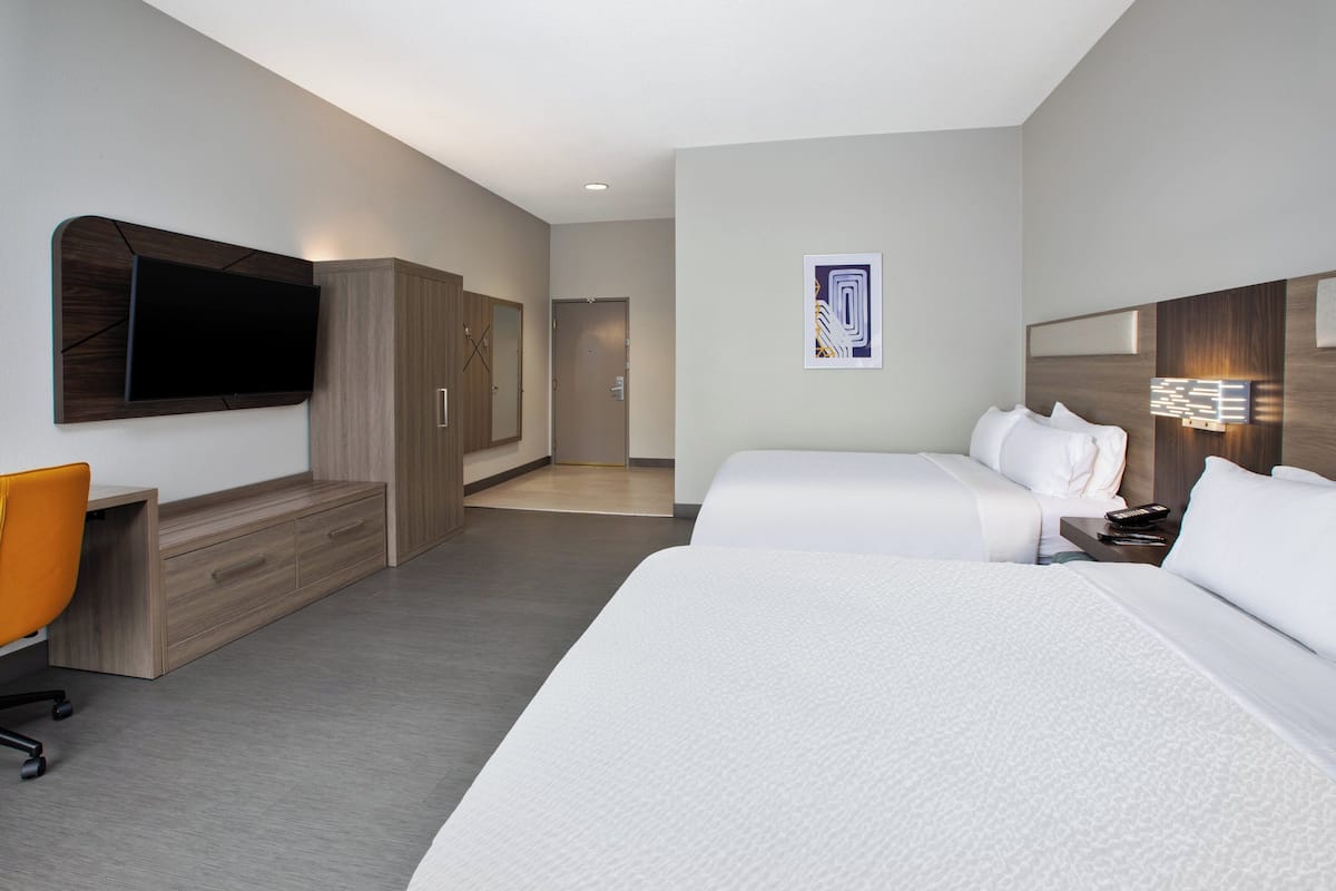 A hotel room with two beds, a desk with a chair, a TV mounted on the wall, and a door leading to another room.