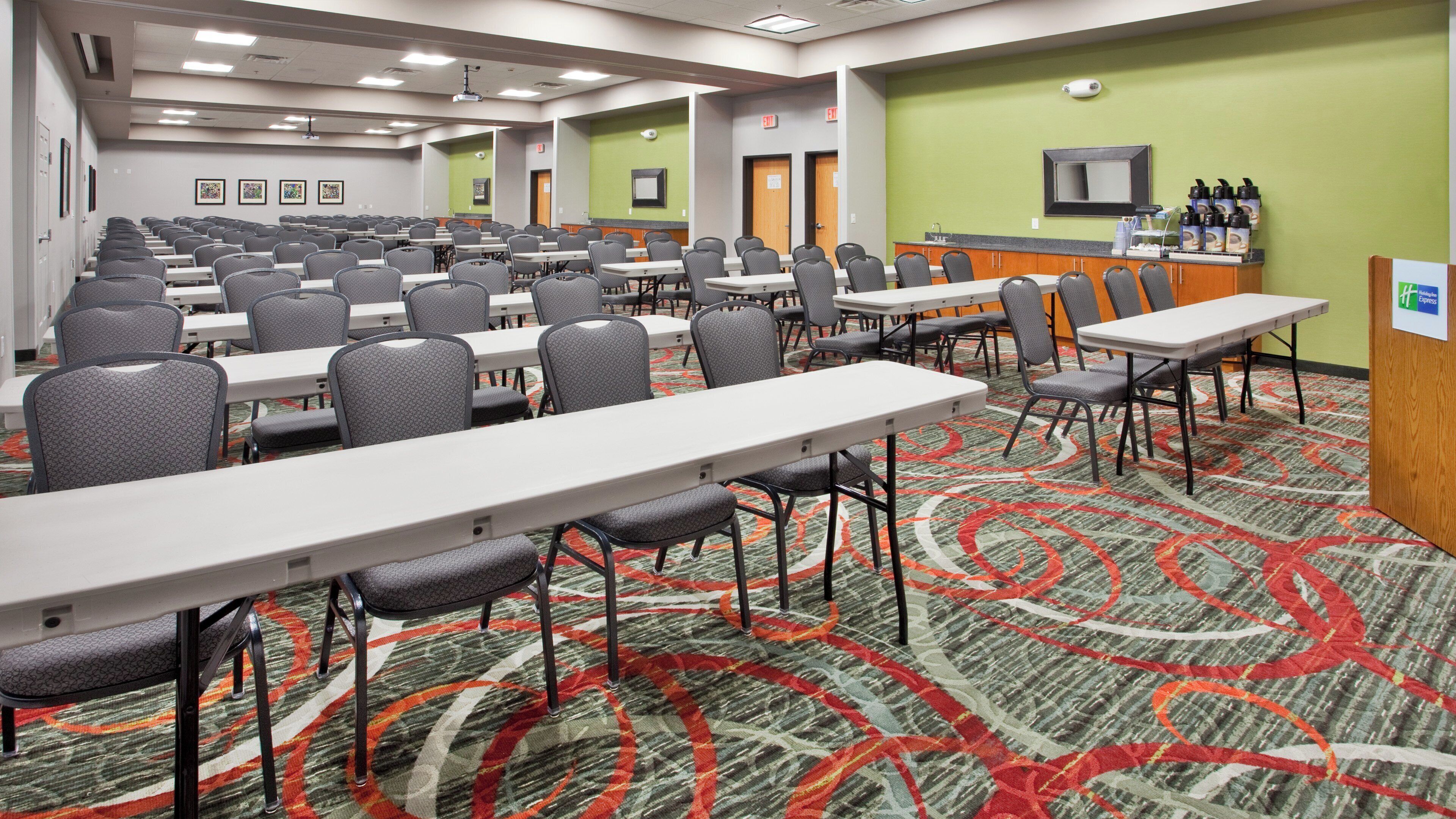 A conference room with rows of tables and chairs, a carpeted floor, and a kitchenette area.