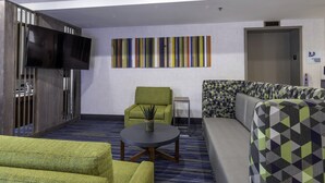 Habitación de hotel con una televisión de pantalla plana montada en una pared de madera, un sillón verde, una mesa redonda con una planta, un sofá estampado y una alfombra a rayas.