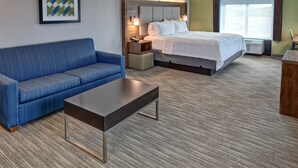 A hotel room with a blue sofa, a bed with white linens, a wooden headboard, a nightstand with a lamp, and a large window. - Holiday Inn Express Hotel & Suites Memphis Southwind by IHG (Memphis)