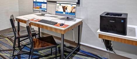 A hotel workspace with two desktop computers, a printer, and a chair with a laptop bag.
