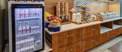 A breakfast bar with a refrigerated display of yogurt and milk, a fruit bowl, cereal, and various breakfast items.