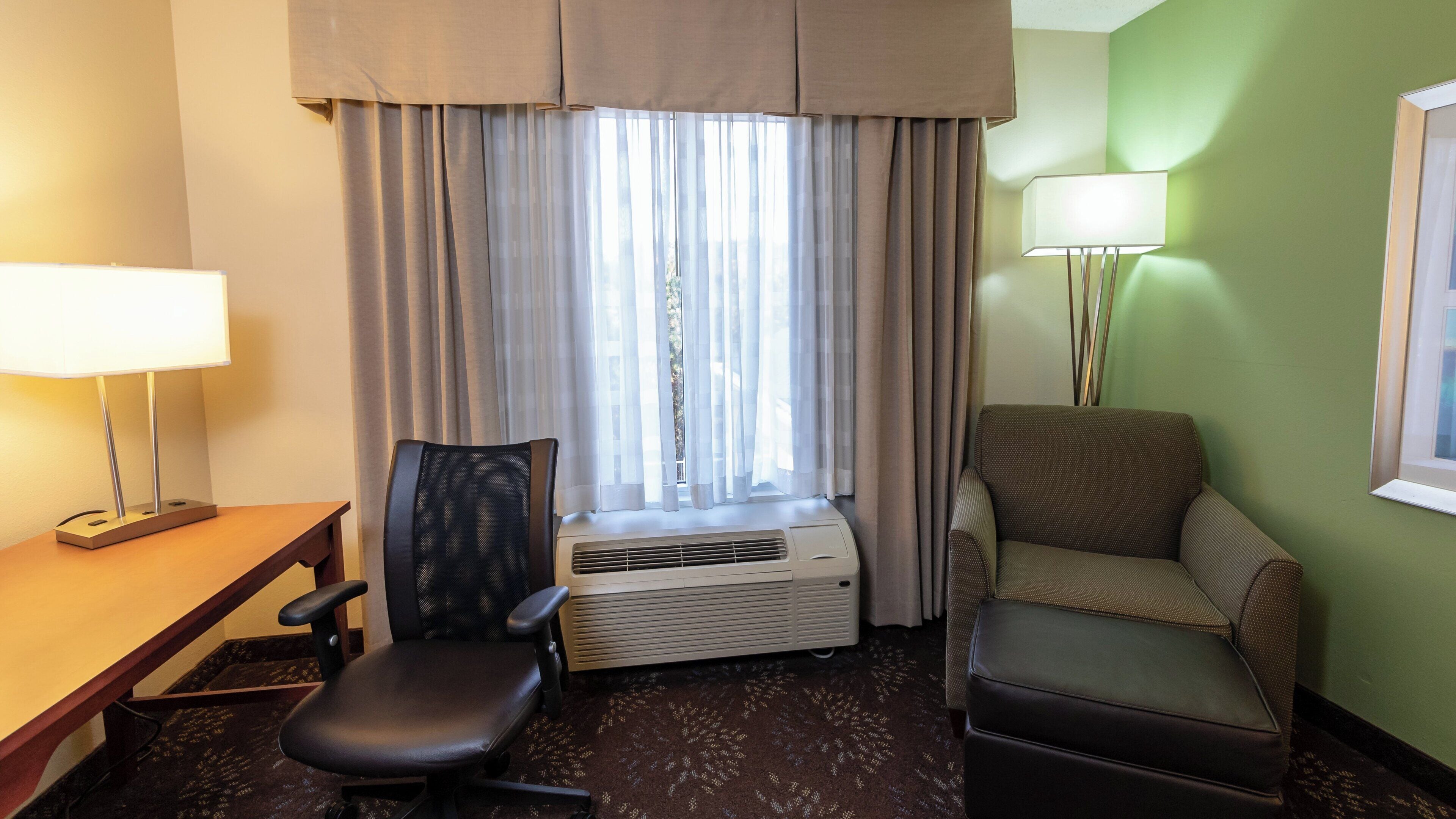 Habitación de hotel con escritorio, dos sillas, aire acondicionado, ventana con cortinas y lámpara.
