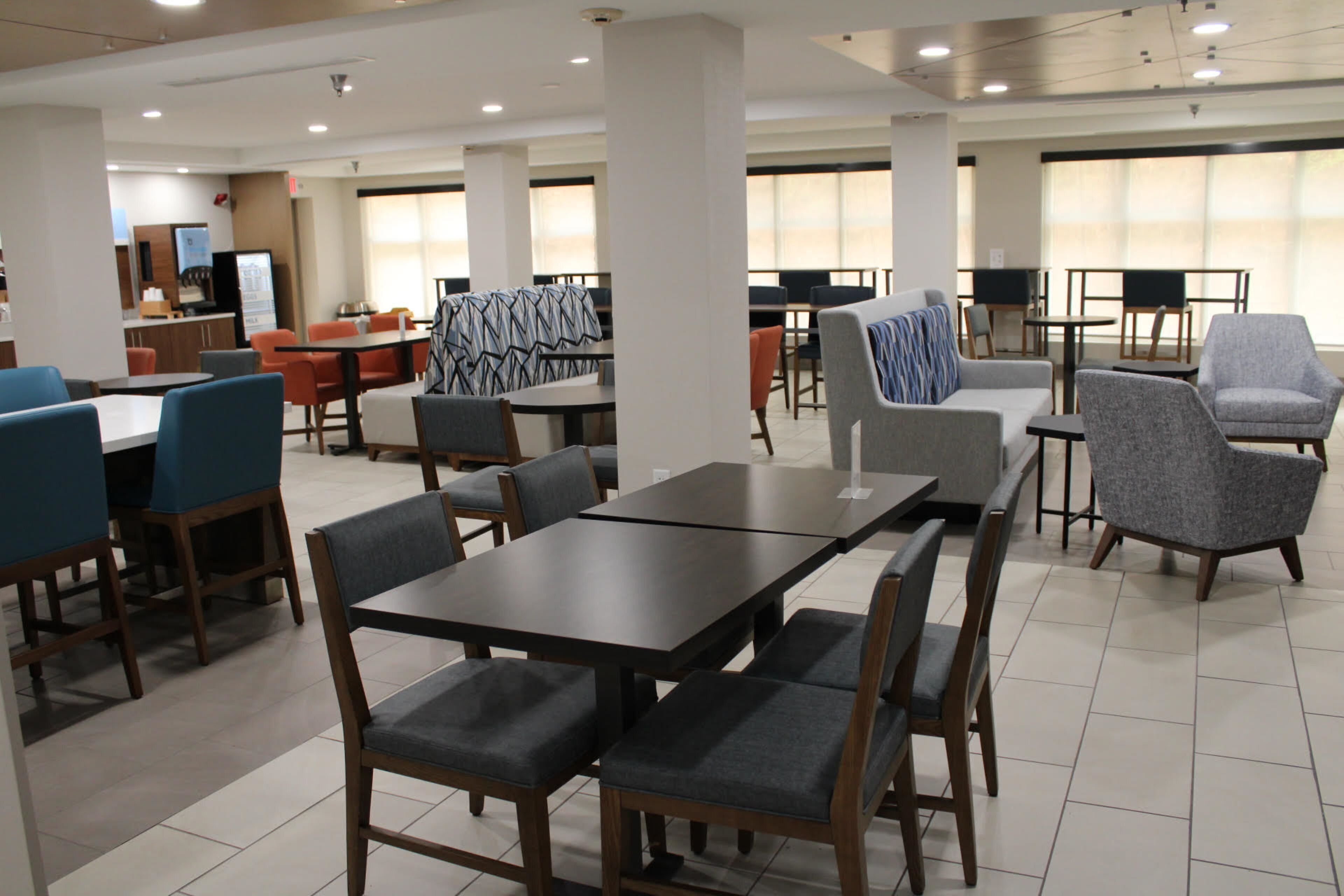A spacious dining area with multiple tables and chairs, a counter with a coffee machine, and large windows providing natural light.